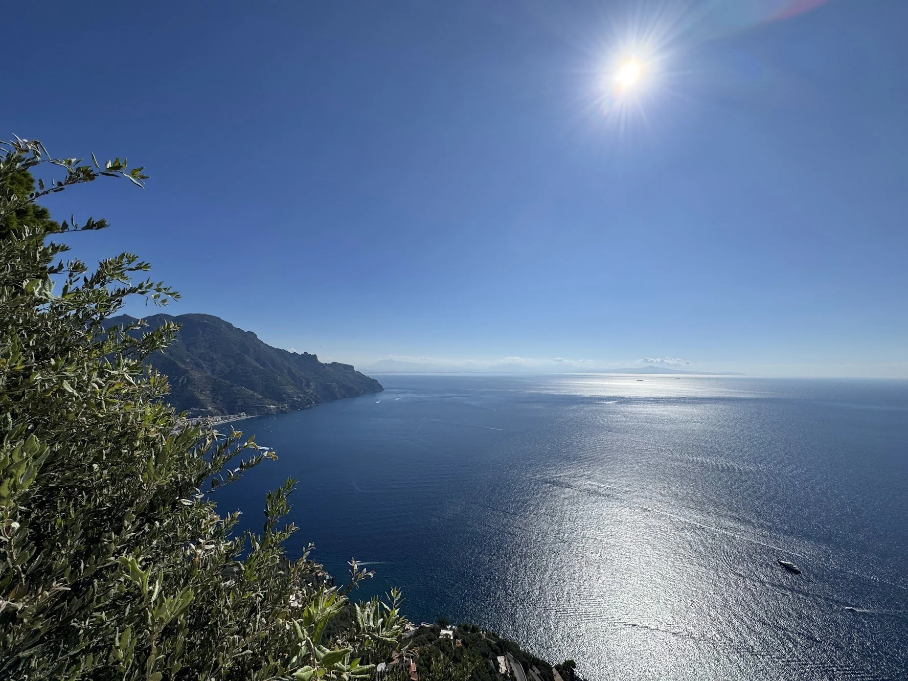 A coastal landscape with a bright sun in a clear blue sky, rugged green hills along the shoreline, and a calm ocean with a small boat in view.