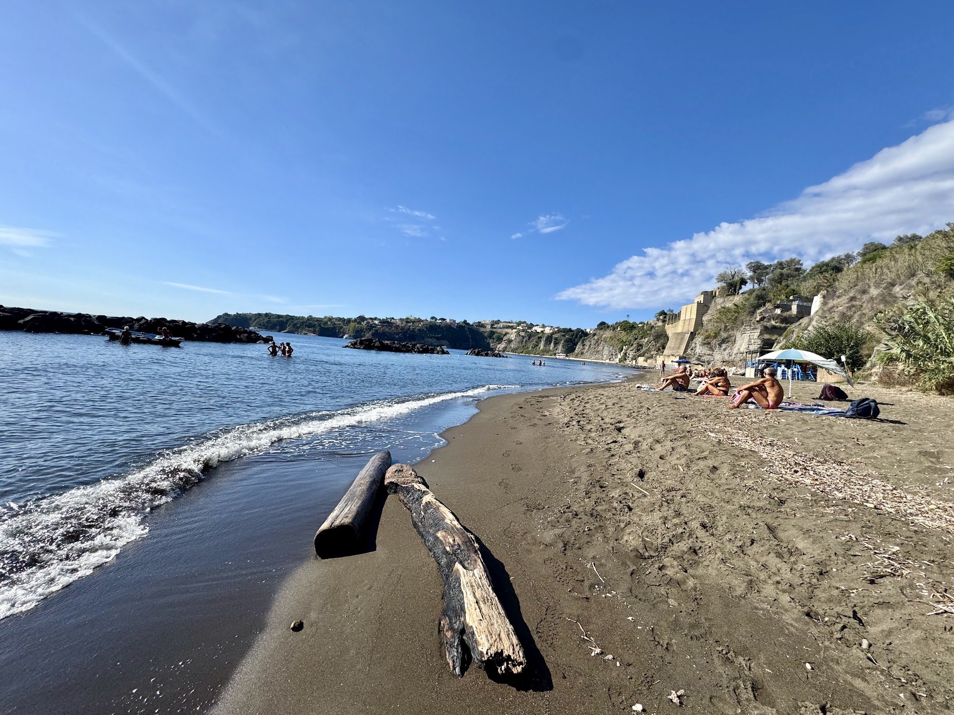 People relaxing on a sandy beach with a picnic umbrella, driftwood, rocky shoreline, and cliffs in the background, under a clear blue sky.