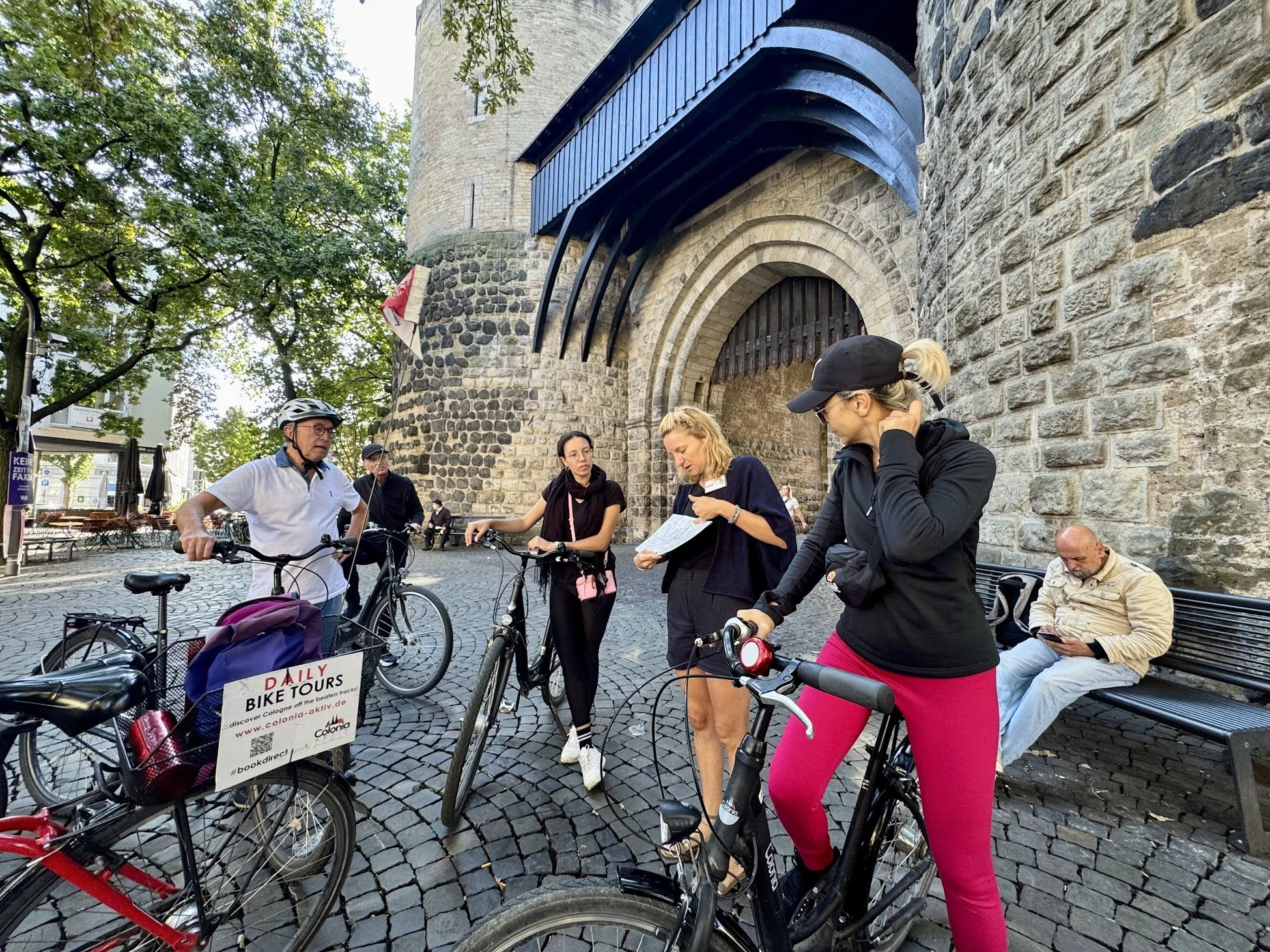 Group of people gathered with bicycles in front of a historic stone building with a tower. Some are looking at maps or their phones, and one person is wearing a helmet. The scene is lively with a mix of men and women.