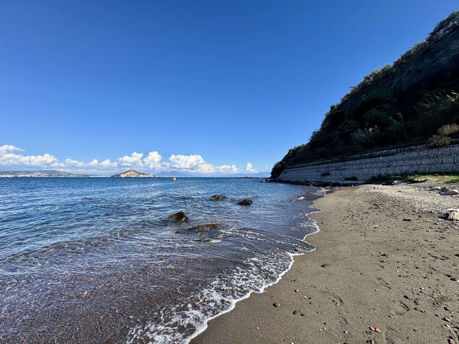 Beach with sandy shoreline, small rocks, calm ocean water, stone wall on hillside, clear blue sky with some clouds, distant land and small island.