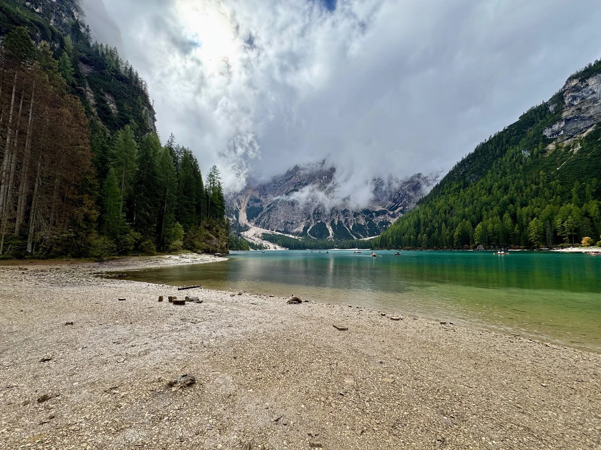 View of a mountain lake surrounded by dense green trees with a rocky mountain in the background, partially covered by clouds and mist.