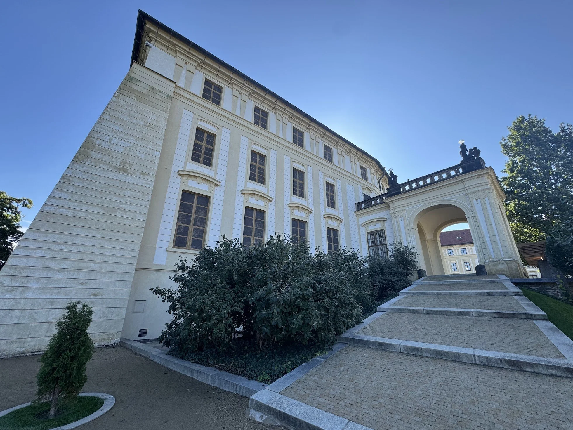 An elegant, historic building with cream-colored walls and multiple tall windows, featuring classical architectural details, a large archway at the entrance, and decorative statues on the rooftop, set against a clear blue sky with some trees in the b
