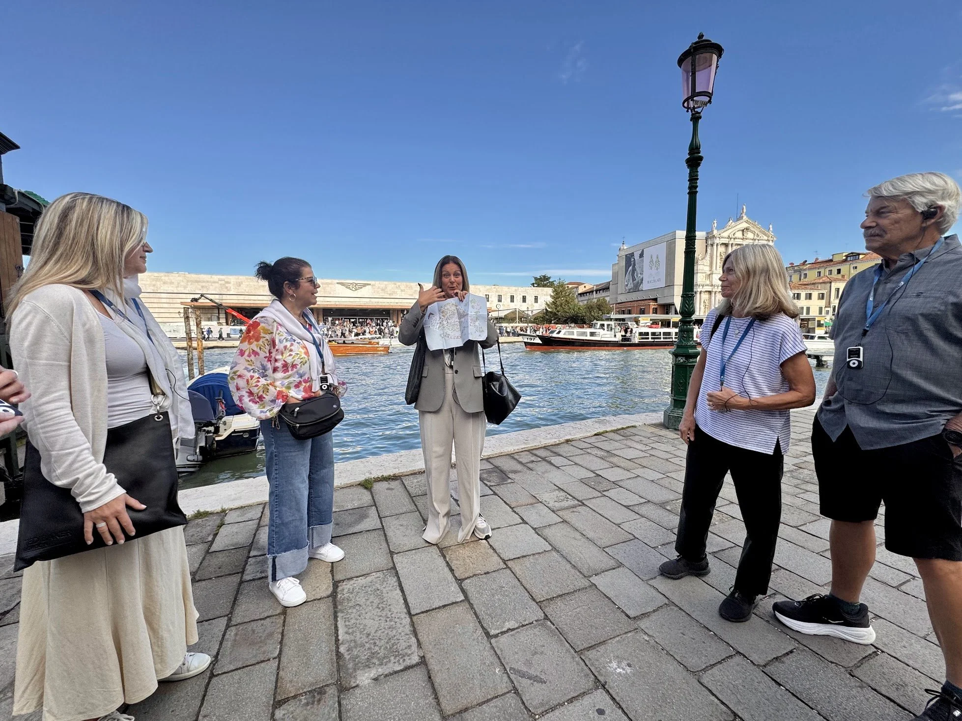 Group of people standing near a river, some holding cameras, with a woman showing a map, historic buildings, and a boat in the background on a sunny day.