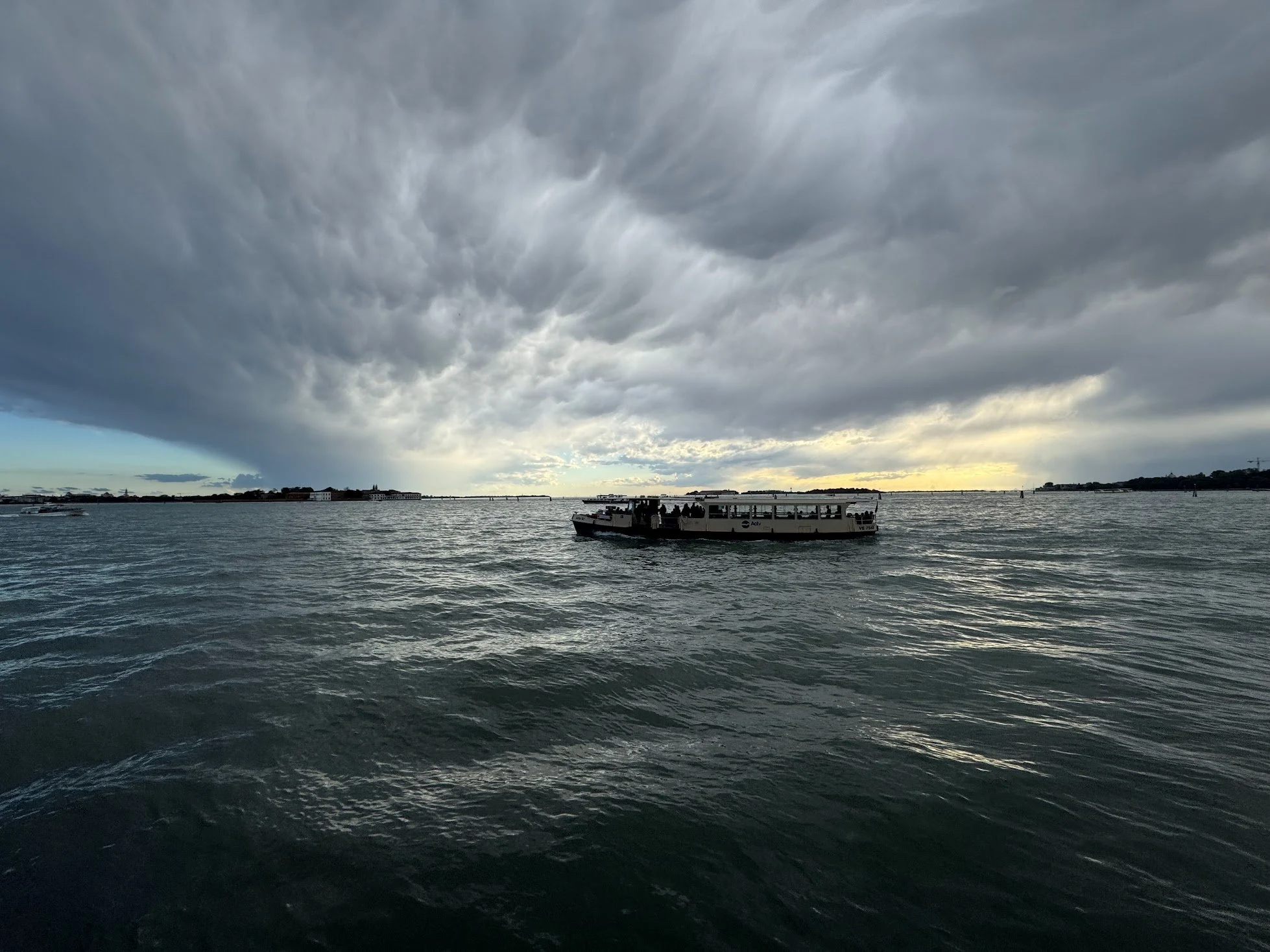 Overcast sky over a body of water with a boat in the foreground and buildings on the distant shoreline.