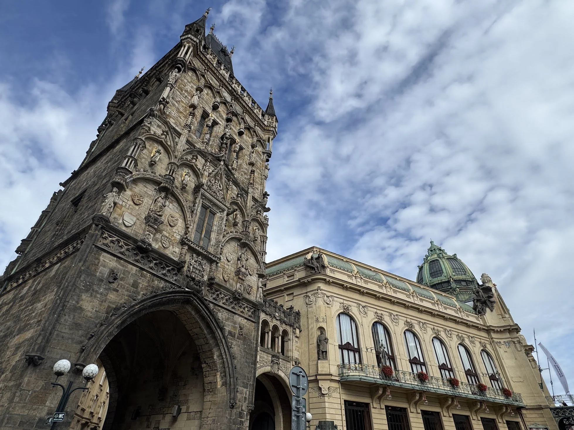A historic European building with ornate architecture, stone statues, and a clock tower topped with a pointed spire, against a cloudy sky.