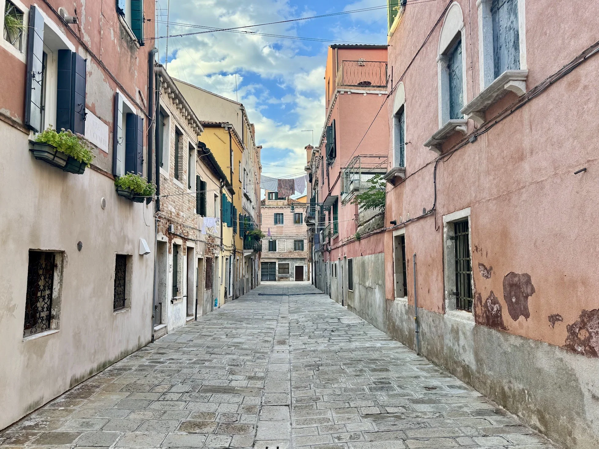 A narrow cobblestone street lined with colorful traditional European buildings with small windows and shutters, laundry hanging on lines above, under a partly cloudy sky.