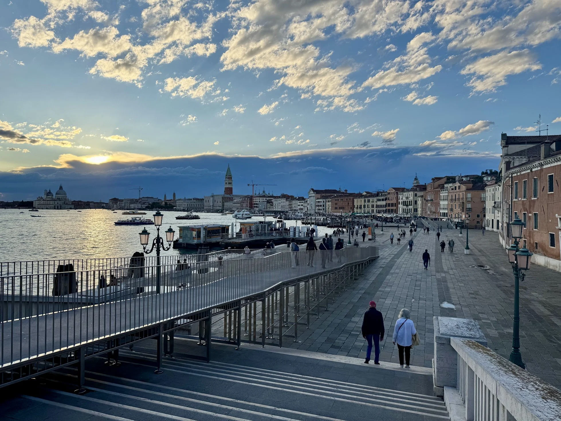 View of a waterfront cityscape at sunset with clouds, boats on the water, and historic European-style buildings along the shoreline.