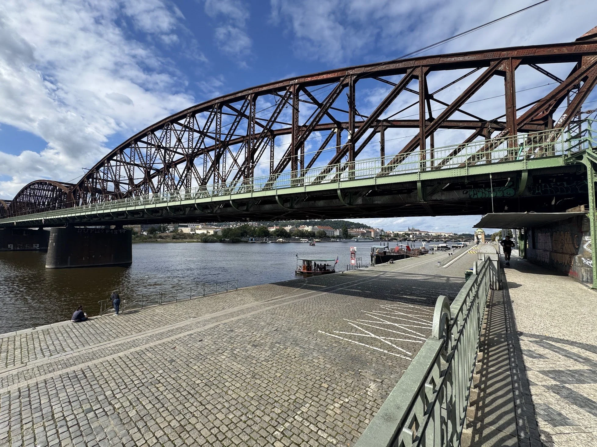 A large steel bridge crossing over a river, with a waterfront promenade below. The sky is partly cloudy with blue patches. On the promenade, a few people are sitting and walking, and there is a small boat on the river.