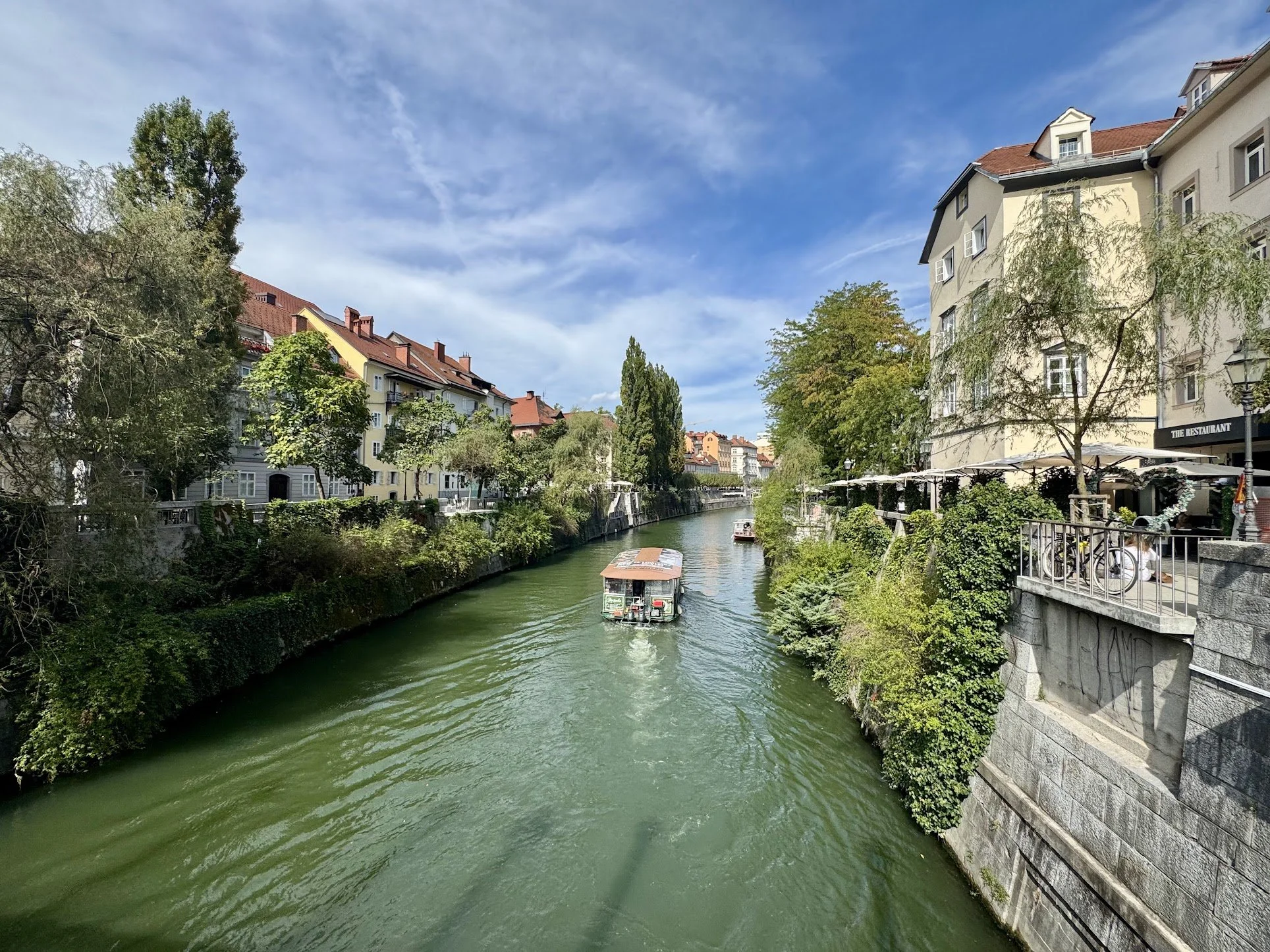 A canal with green water flowing through a city, lined with trees and buildings on both sides. There are boats on the canal and outdoor seating with umbrellas along the right side.