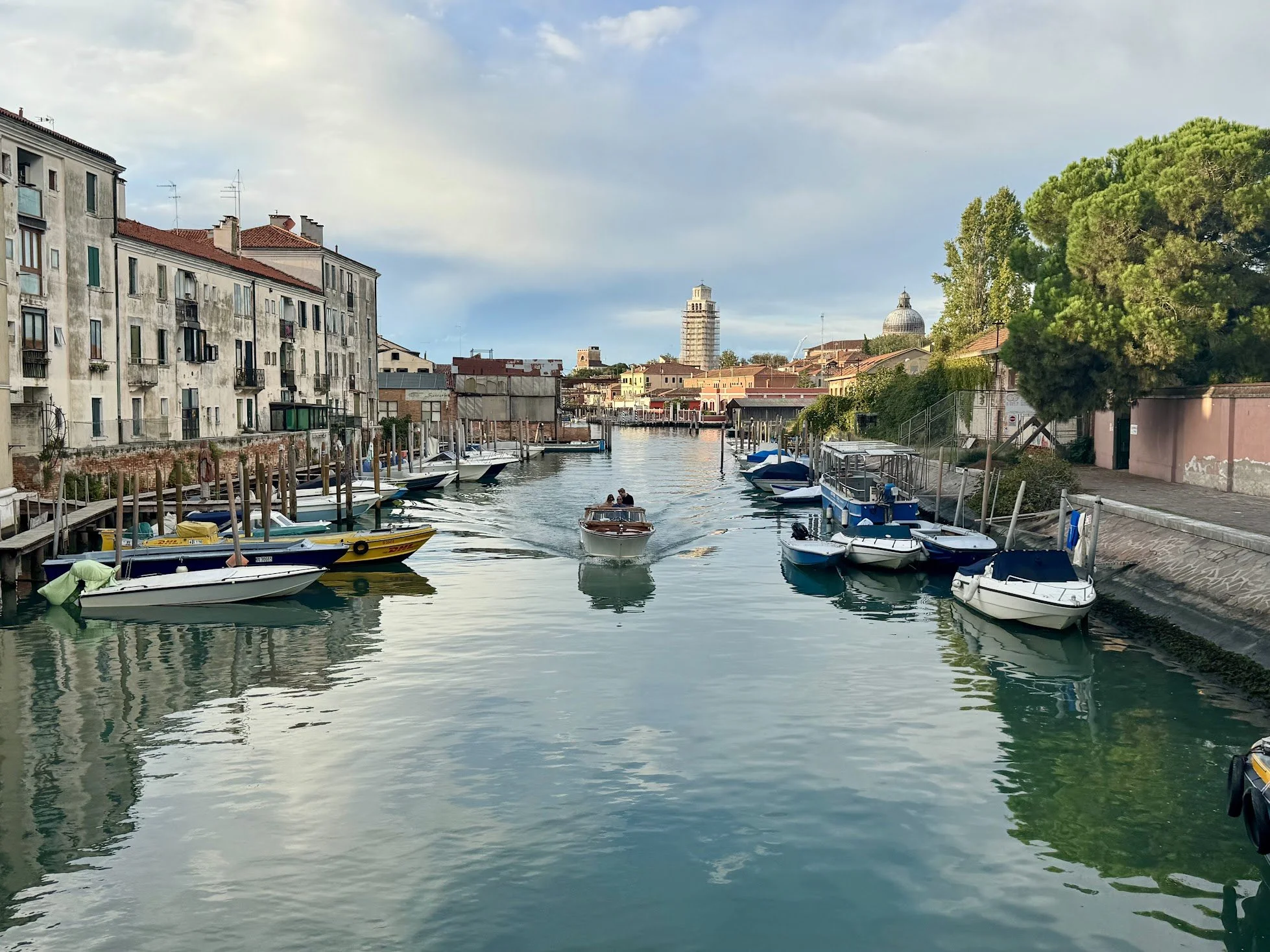 View of a canal with boats docked on the sides, and a small boat with two people sailing in the water, with buildings and greenery in the background under a partly cloudy sky.