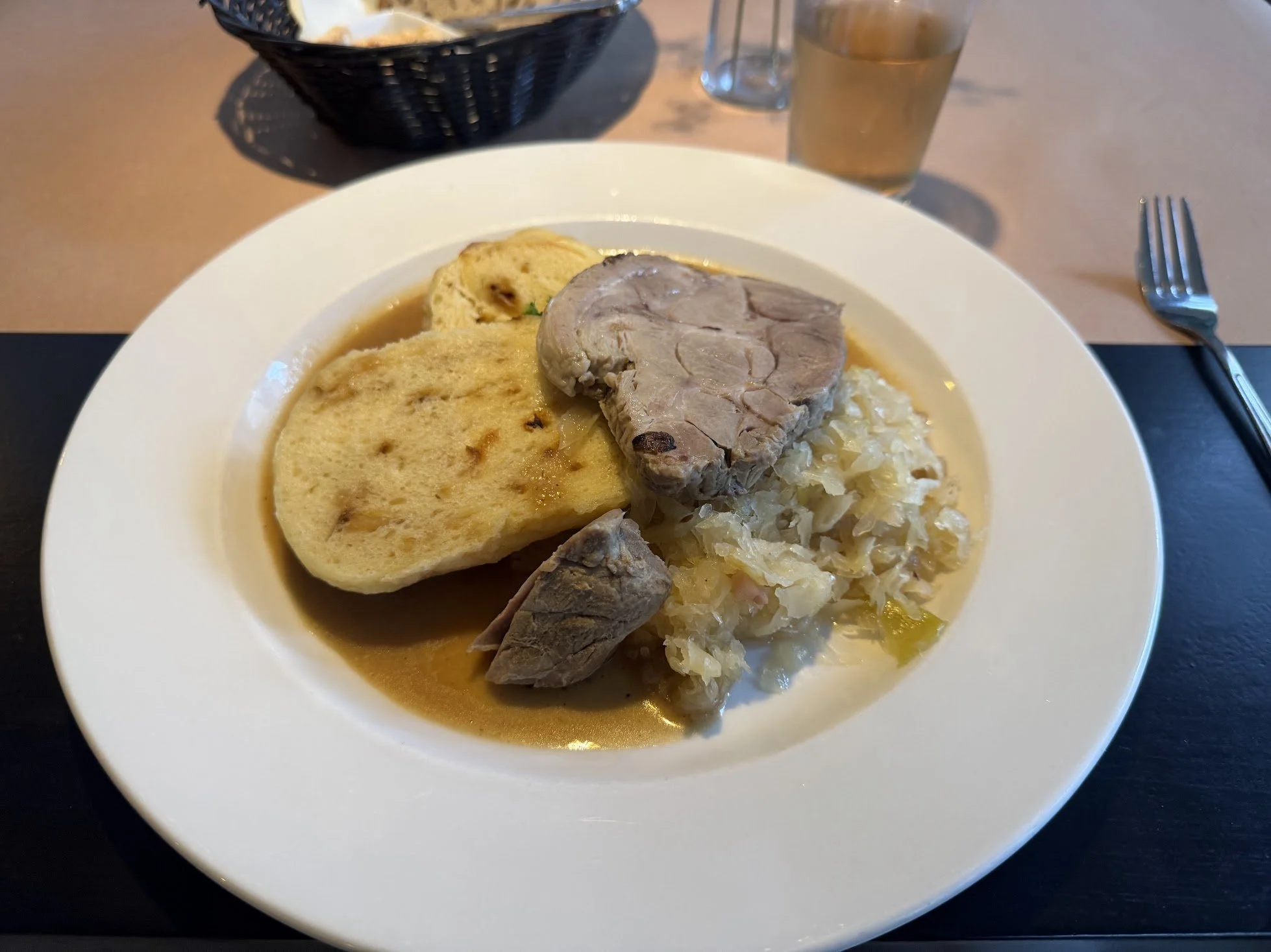 A white plate with mashed potatoes, a slice of meatloaf, a bread roll, and a small piece of ham. In the background, there is a glass of iced tea, a small glass of water, and a black basket of bread rolls on a wooden table.