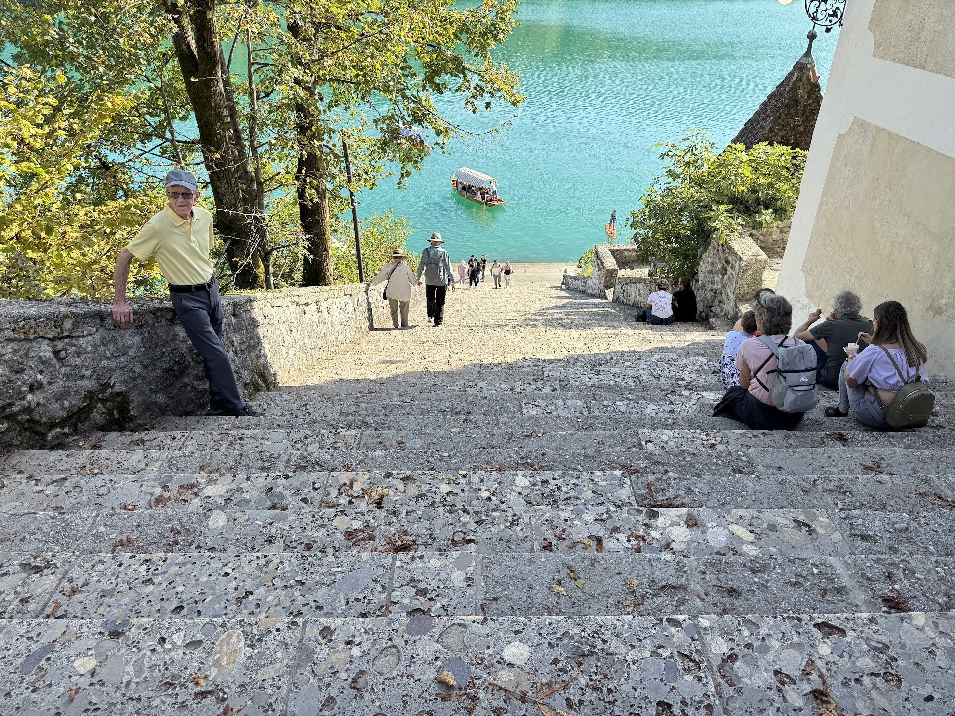 People walking down a stone staircase leading to a lake with a boat and paddleboard on the water. There are trees and buildings nearby.