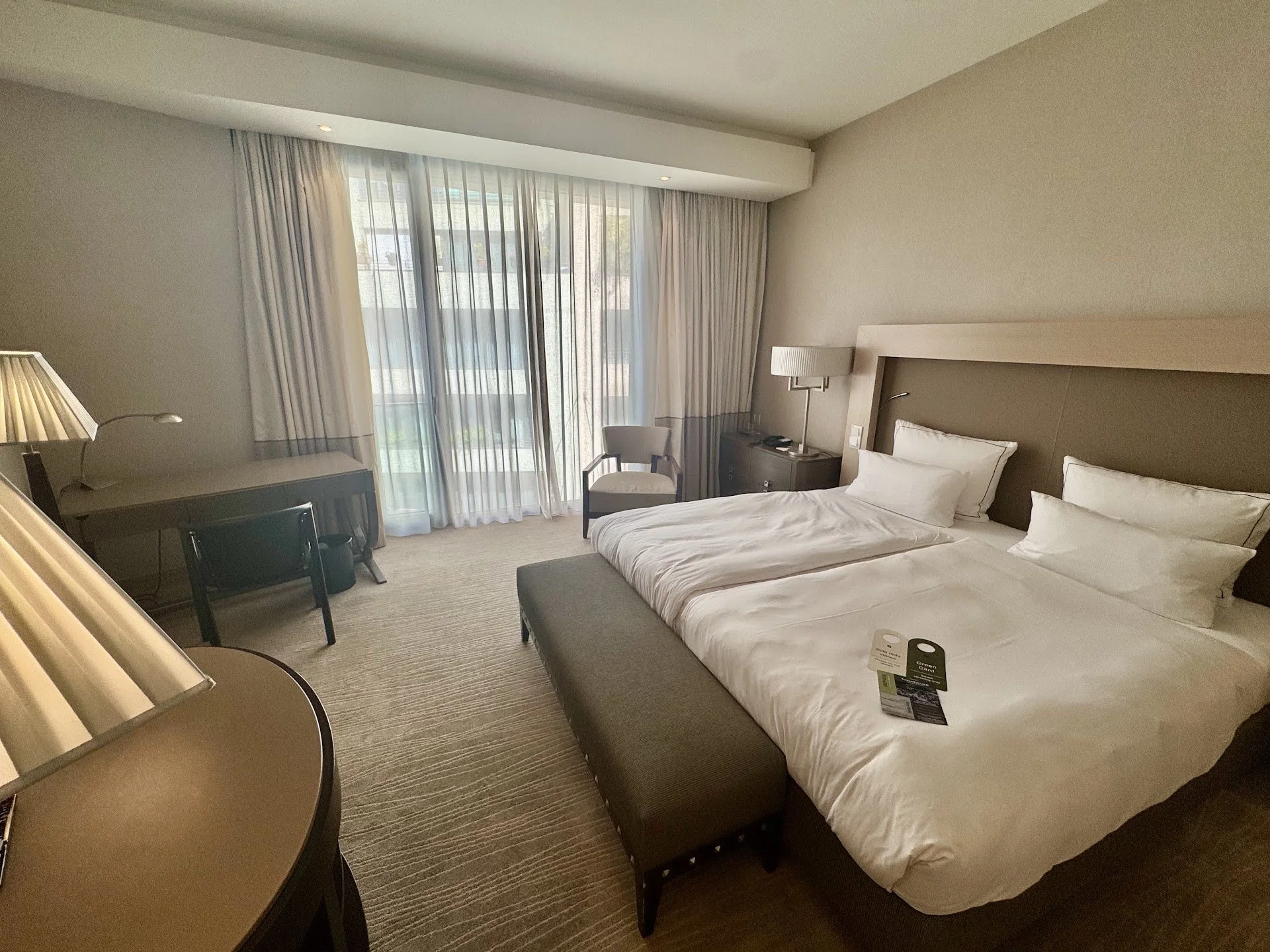 A hotel room with a large bed, bedside tables, lamps, a chair, and a window with sheer curtains revealing a balcony.