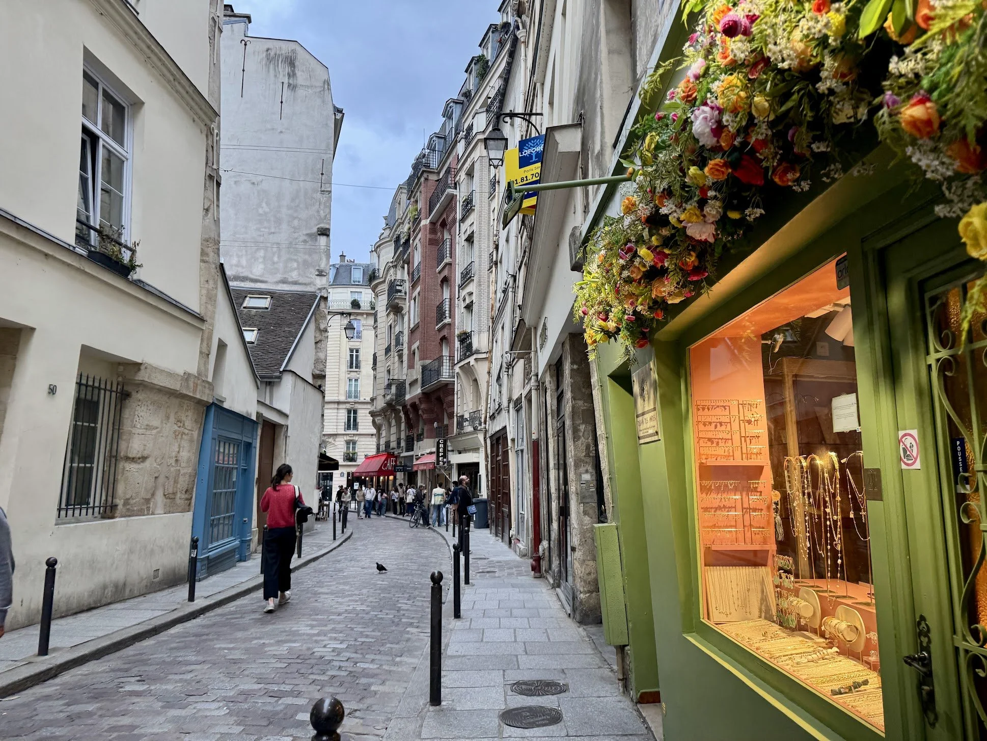 A narrow street in a European city with cobblestone pavement, lined with tall, multi-story buildings with balconies. On the right, a jewelry store with a bright green facade and a window display illuminated with orange light. Several people are walki