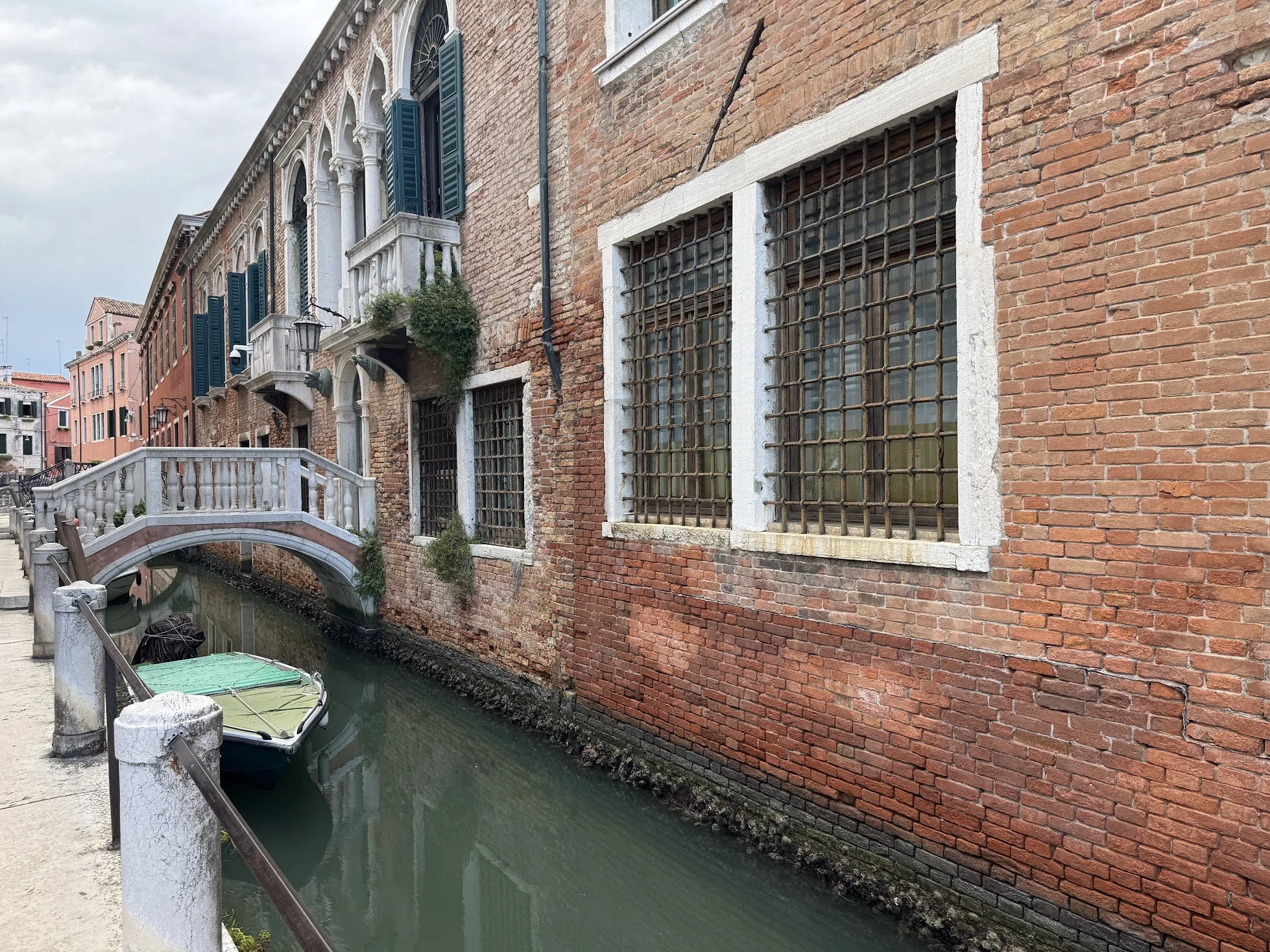 A canal with a small boat floating beside a brick building with barred windows and green shutters in Venice, Italy. A small bridge crosses over the canal.