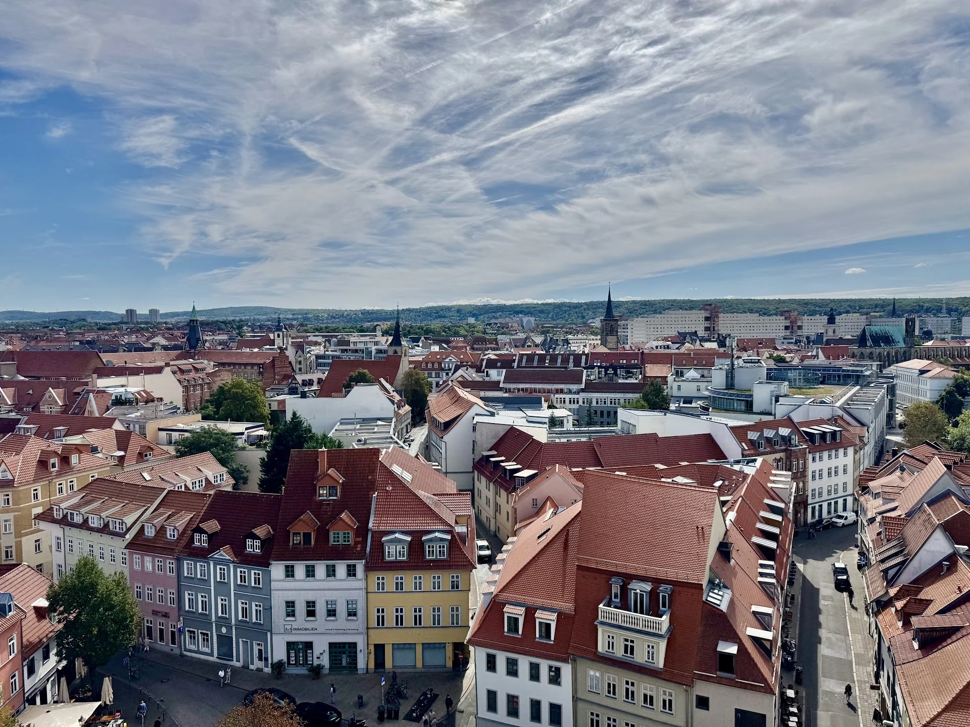 A cityscape with tightly packed buildings and red-tiled roofs, under a partly cloudy sky with wispy clouds.