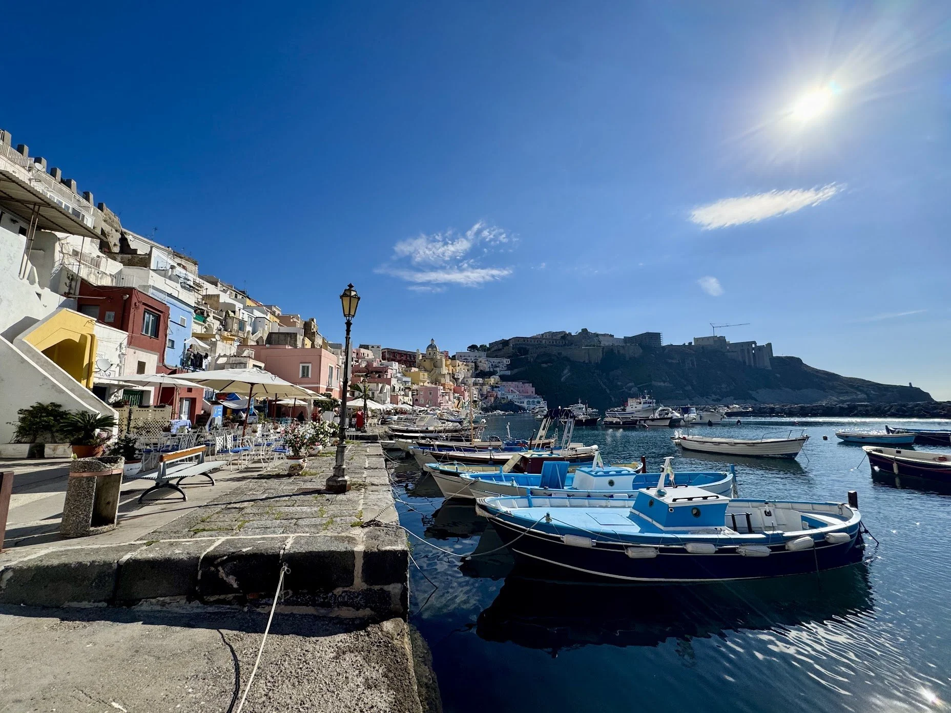 A harbor with several small boats docked, colorful buildings on a hillside, a lighthouse, and fortress ruins in the background under a bright blue sky with the sun shining.