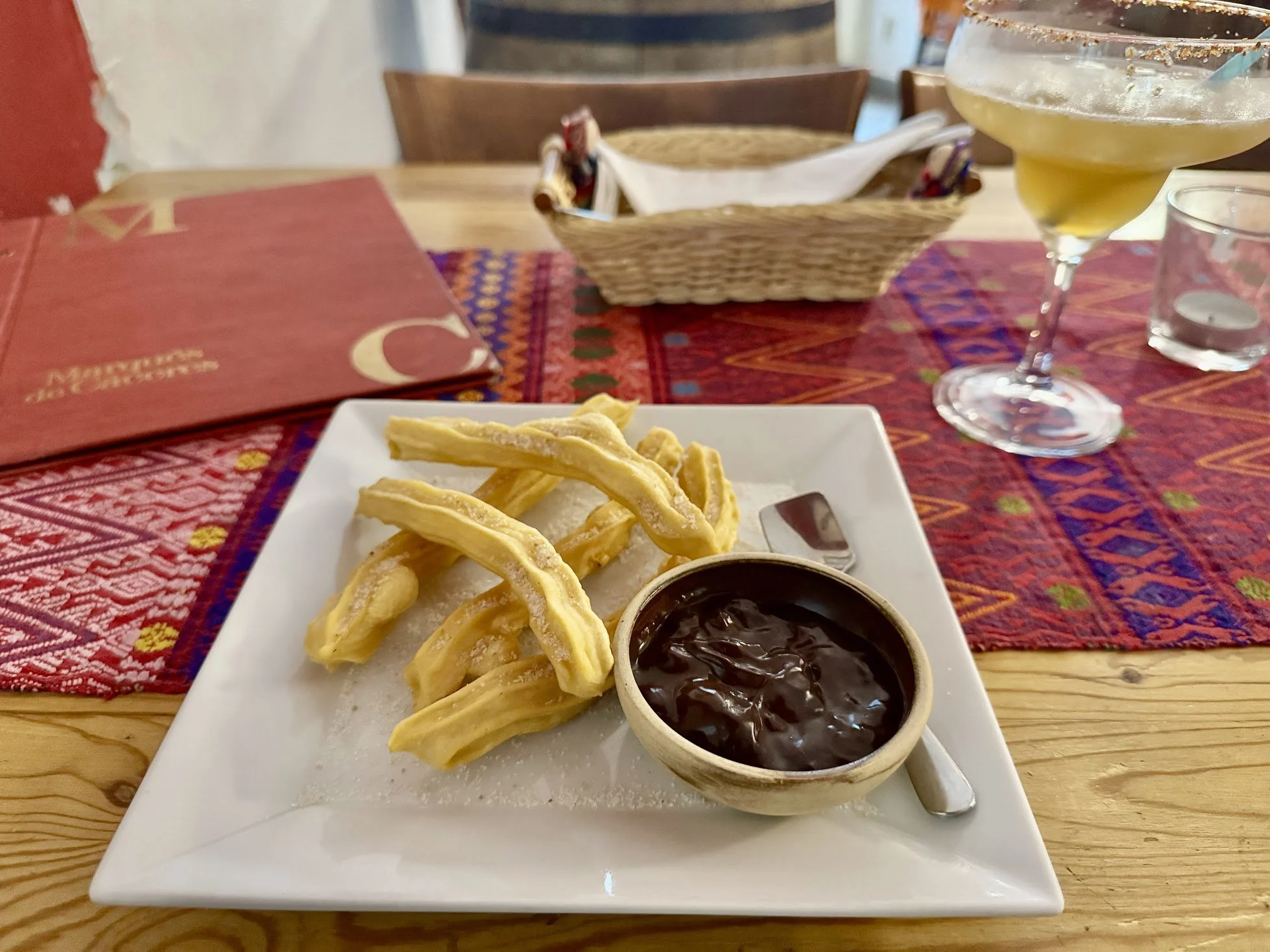 A white square plate with fried churros and a cup of thick chocolate sauce on a wooden table. In the background, there is a colorful tablecloth, a basket with napkins, a cocktail glass with a yellow drink, and a glass of water.
