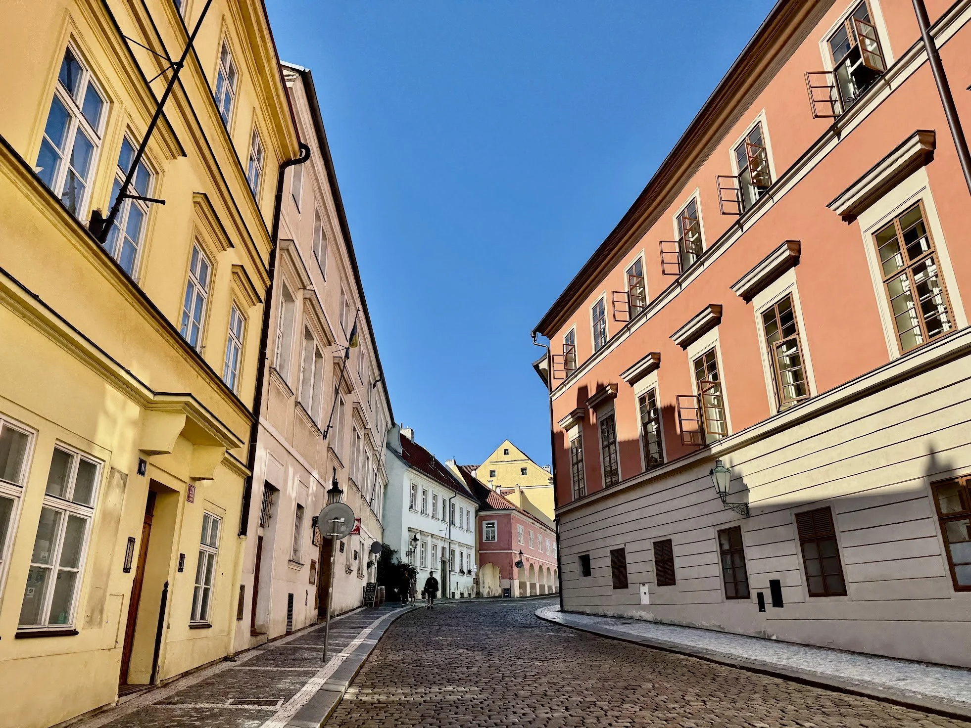 A narrow European street with cobblestone pavement, flanked by pastel-colored buildings with large windows and small balconies, under a clear blue sky.