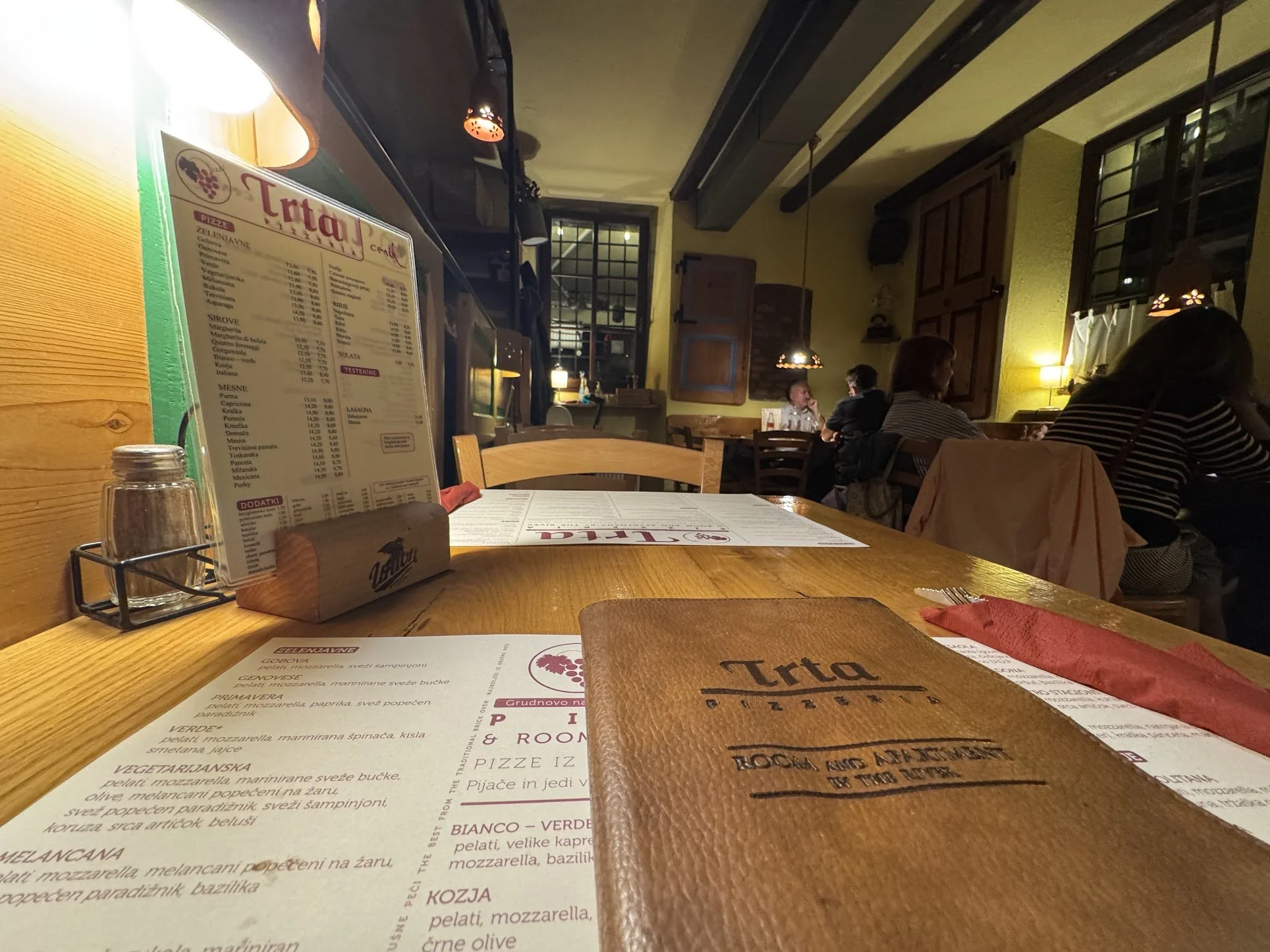 The interior of a cozy Italian restaurant with wooden tables, warm lighting, and a few groups of patrons dining. Menus are on the tables, and there is a mix of modern and rustic decor, with visible windows and wooden beams.