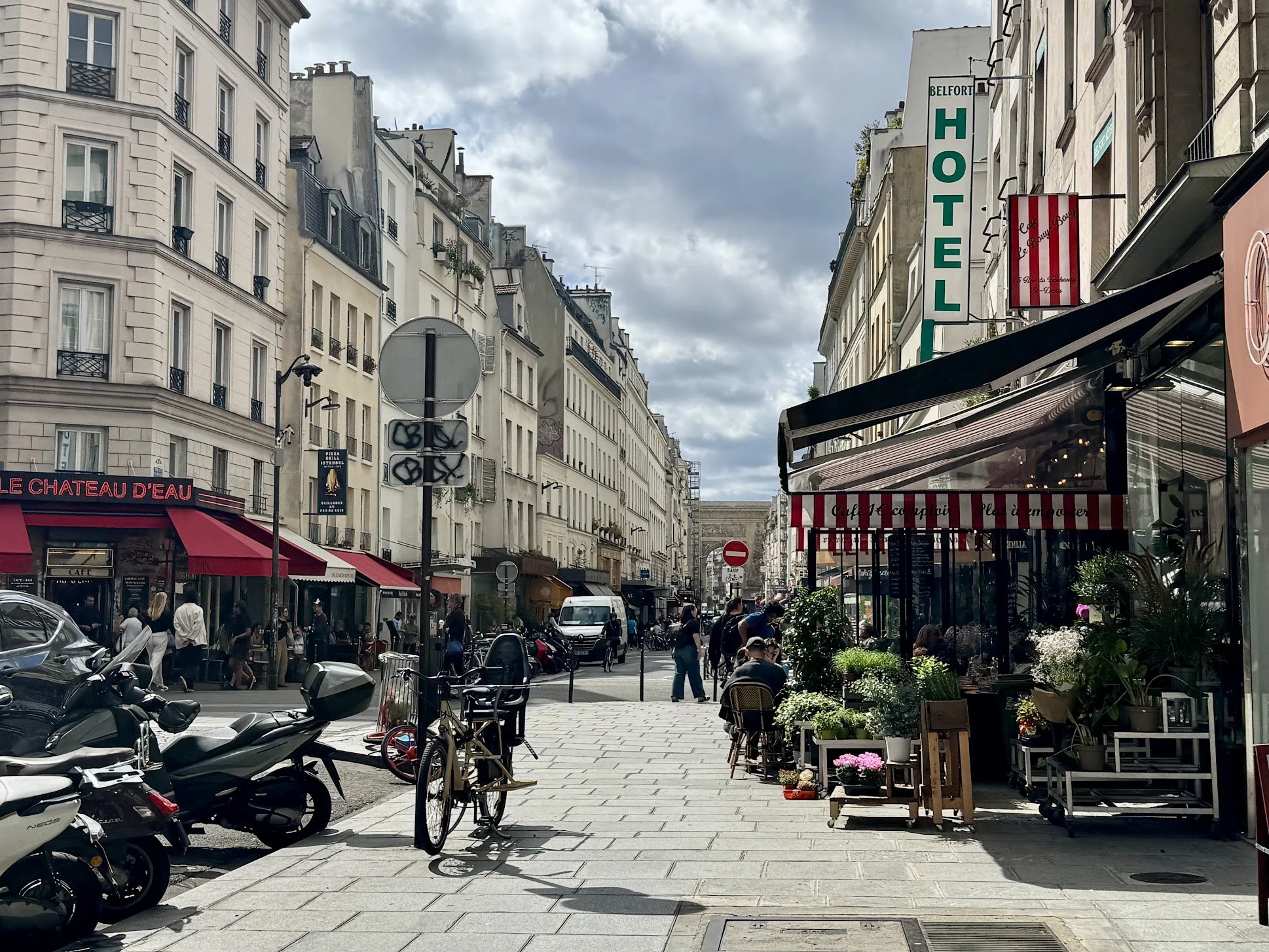 Paris street scene with buildings, outdoor cafes, parked scooters, a bicycle, and pedestrians, under cloudy skies.