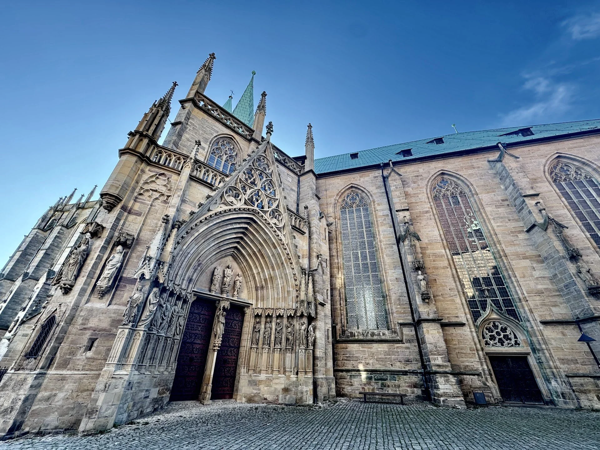 Facade of a Gothic cathedral with detailed stone carvings around the arched entrance, tall stained-glass windows, and green spires against a blue sky.