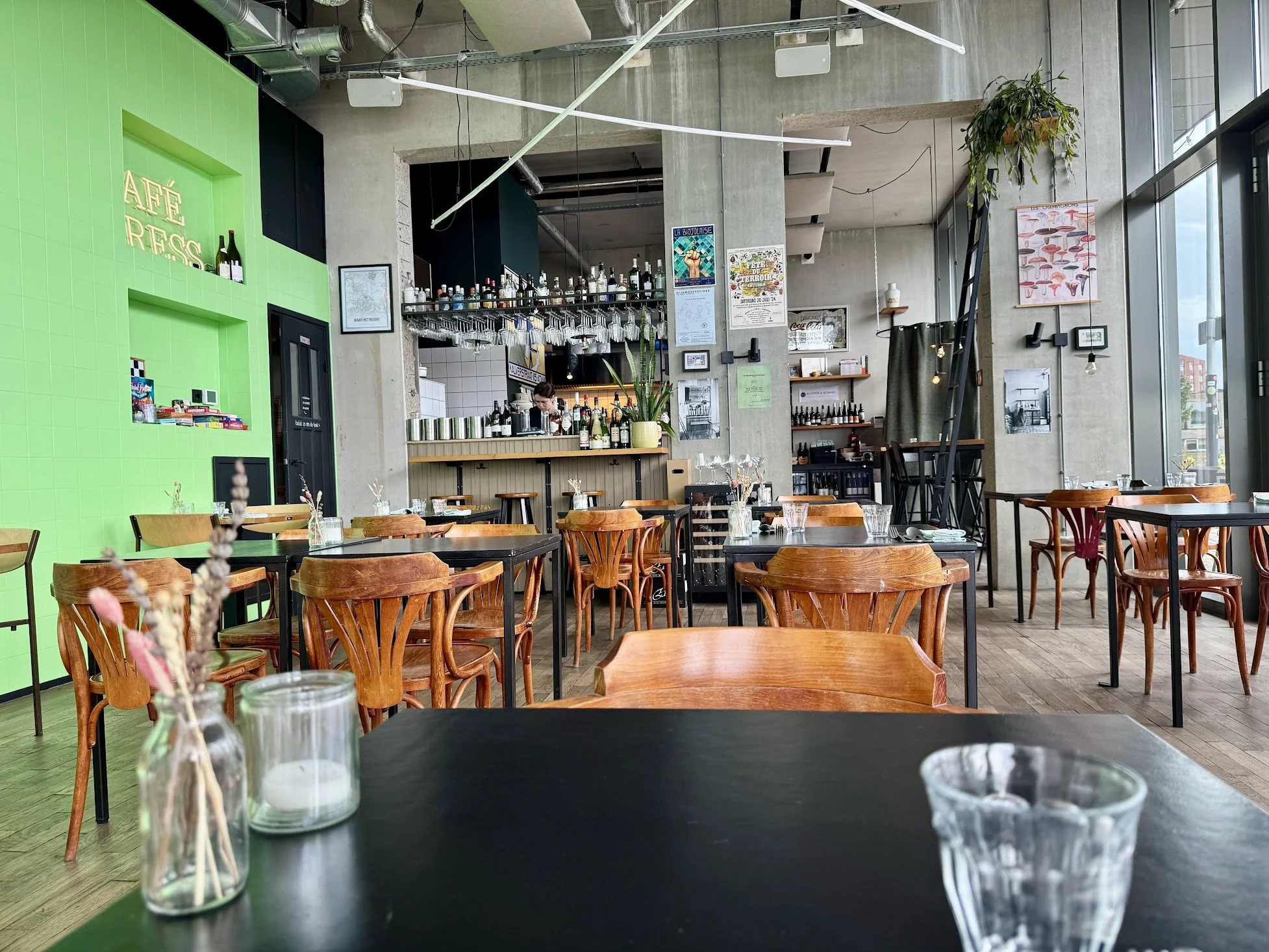 Interior of a modern cafe with wooden tables and chairs, a bar area with bottles and wine glasses, and a large window letting in natural light. Decor includes plants, artwork, and hanging lights.