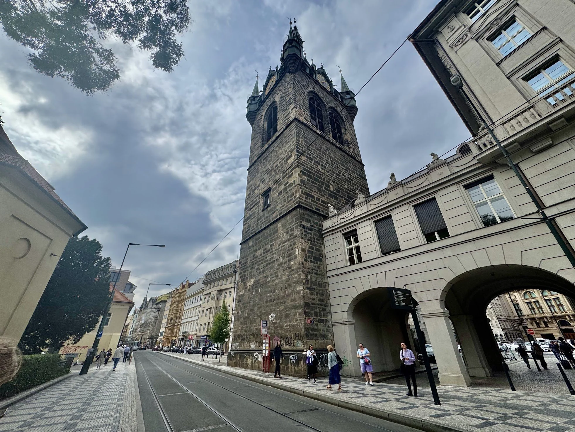 A tall stone tower with a clock, topped with spires and a weather vane, standing on a busy city street with pedestrians walking along the sidewalk, surrounded by historic buildings and trees.