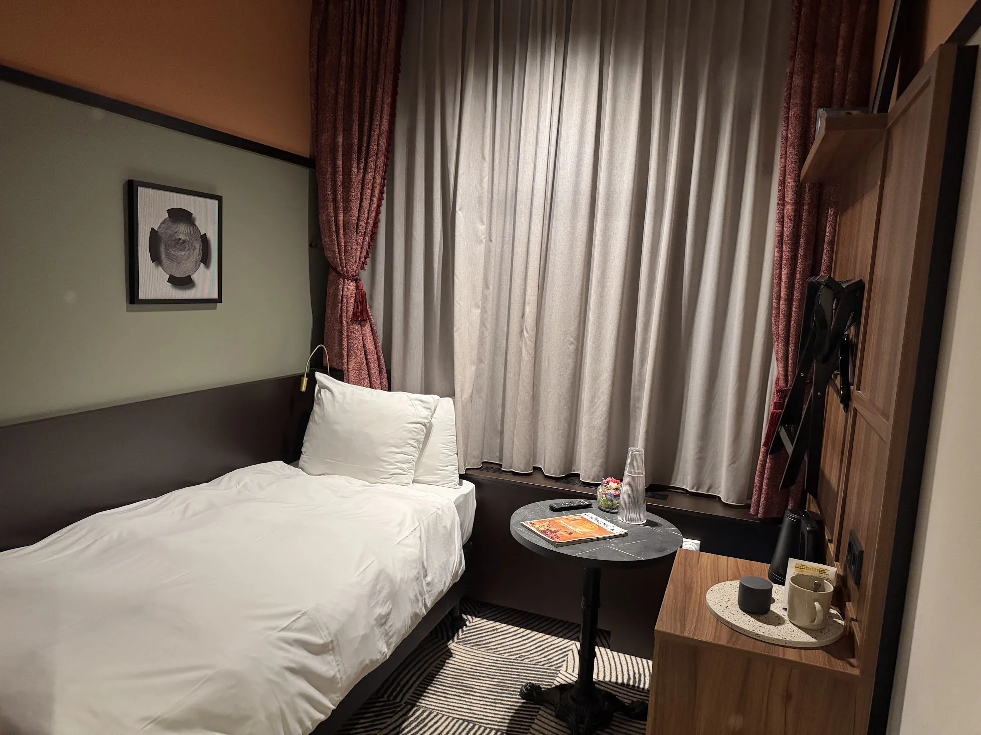 A hotel room with a bed, pillows, nightstand, curtains, artwork on the wall, and a small round table with a magazine, glass vase, and mugs.