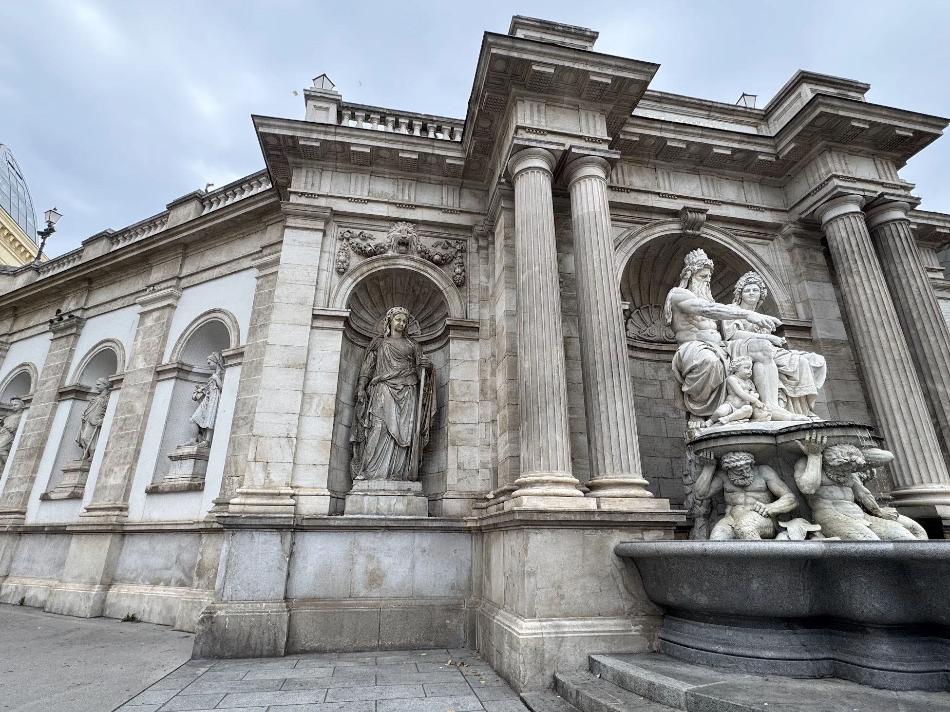 Facade of a classical building with large stone columns, detailed sculptures, and decorative architectural elements, including statues and a fountain in the foreground.