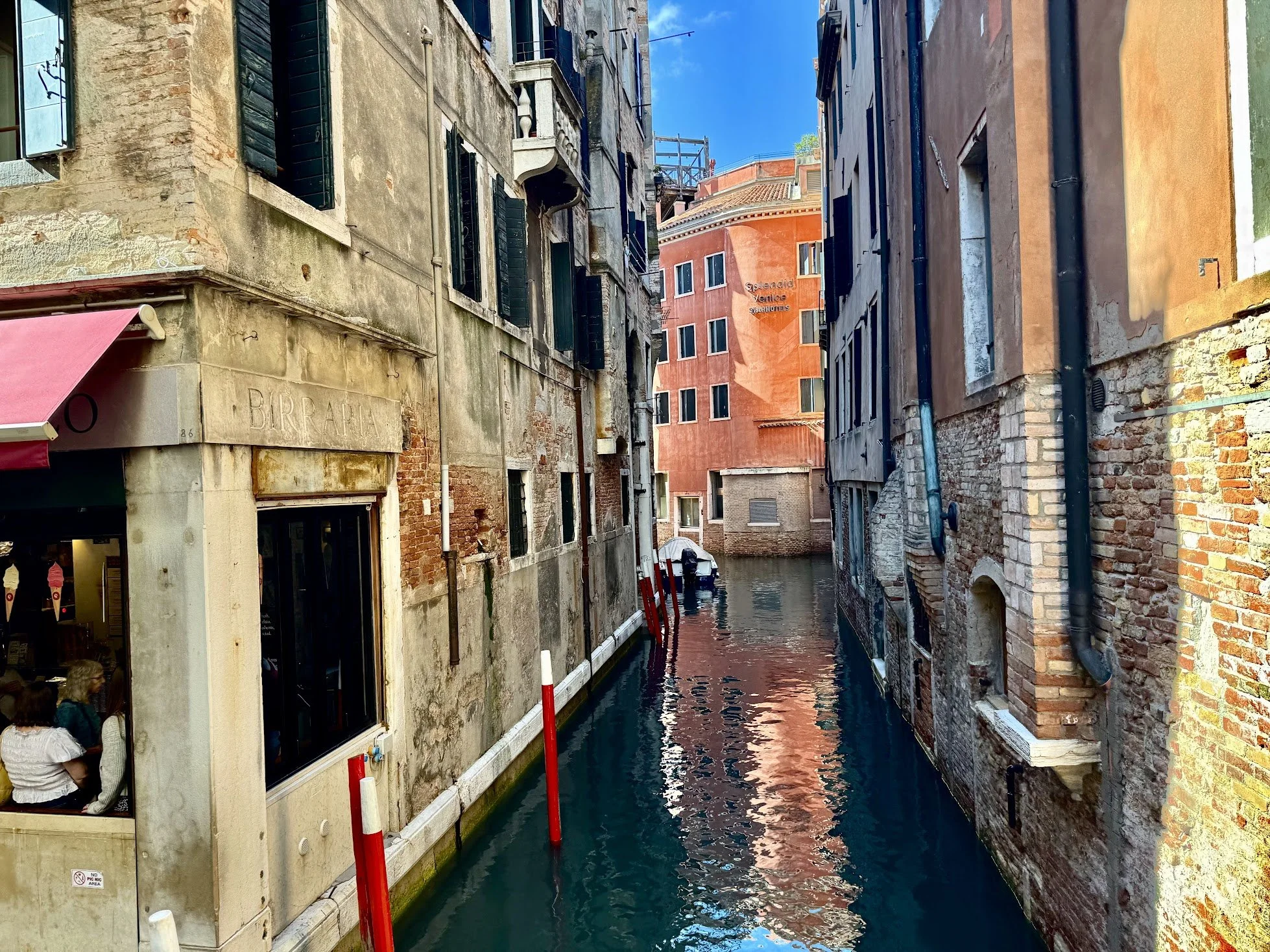 A narrow canal surrounded by old, weathered buildings with colorful facades and windows with green shutters, with a balcony and clothesline visible on some buildings. The water in the canal reflects the buildings and the blue sky with some clouds.