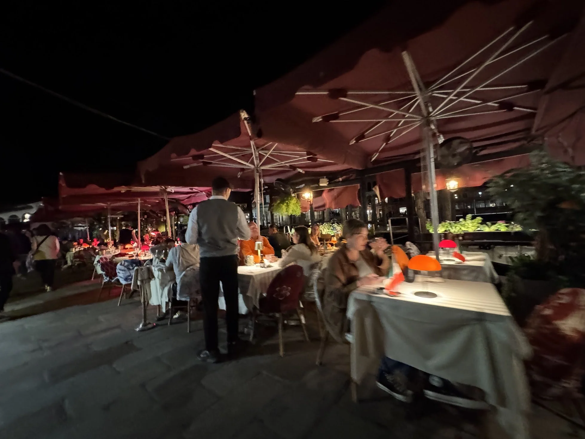 Nighttime outdoor restaurant scene with tables and chairs, patio umbrellas, people dining, and string lights.
