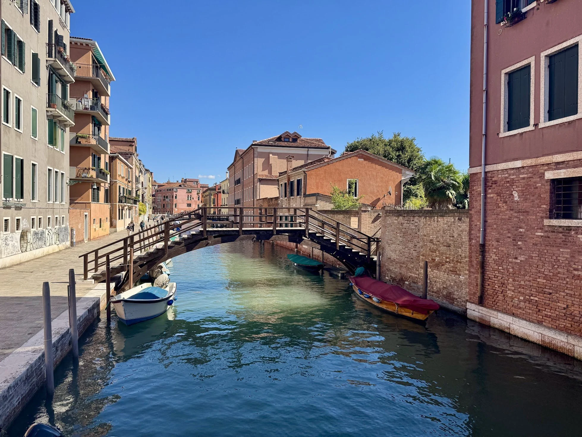 A canal with small boats moored along the sides, surrounded by colorful buildings and a metal bridge, under a clear blue sky.