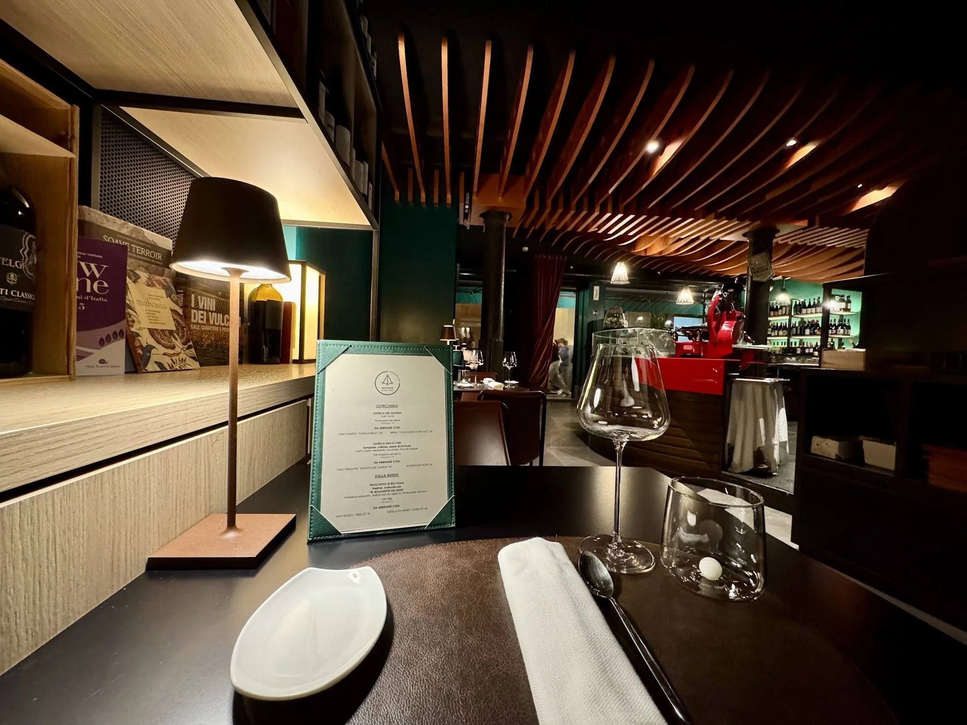 A cozy restaurant interior with wooden ceiling beams, tables set with wine glasses, napkins, and utensils, a menu on the table, and shelves with bottles in the background.