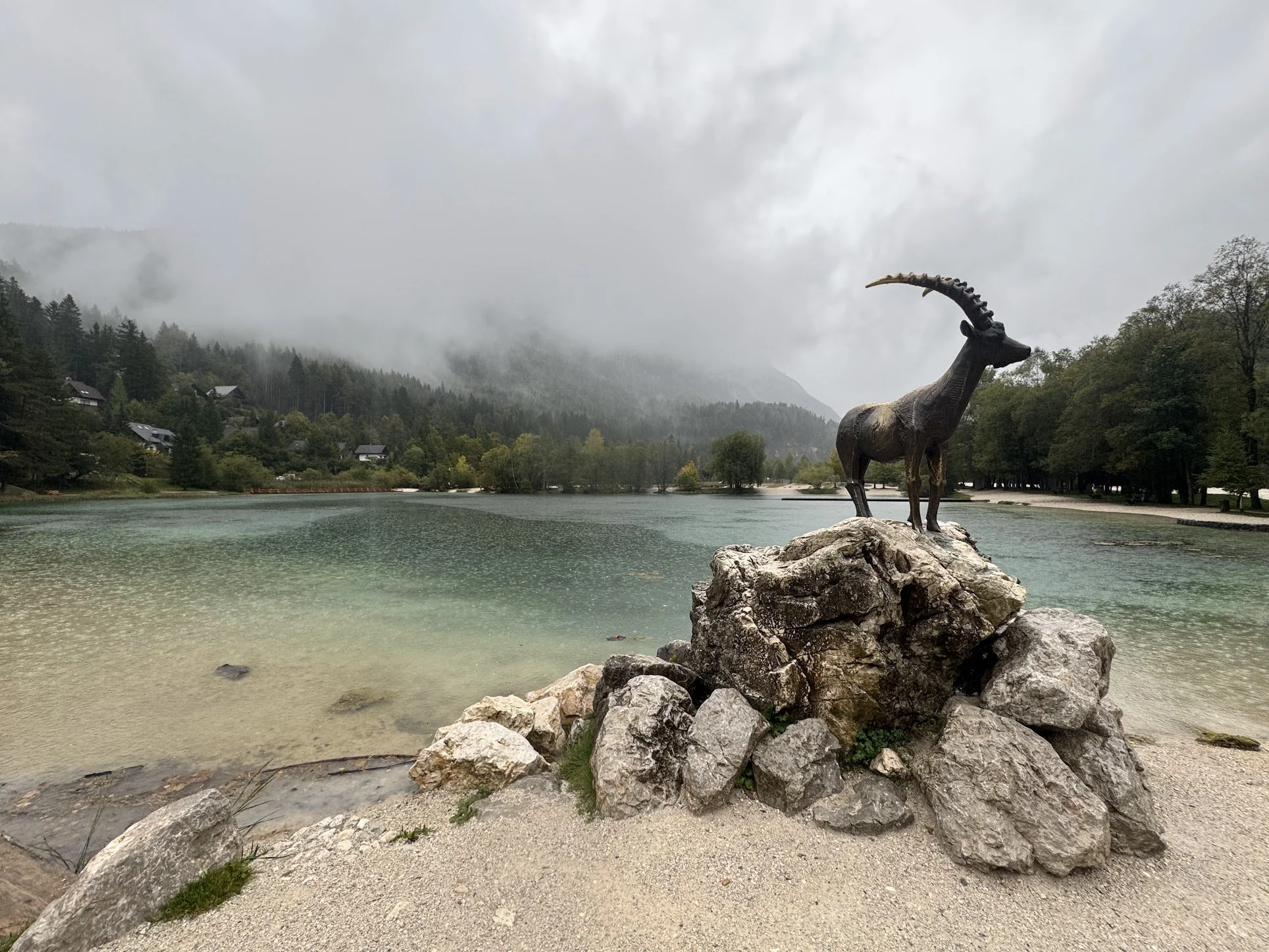 A bronze sculpture of a goat with long, curved horns stands on a pile of rocks by a lakeshore. Surrounding the lake are trees and mountains shrouded in mist and fog, with an overcast sky.