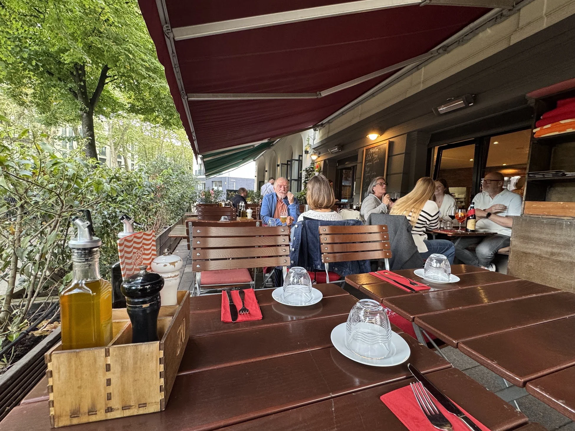 Outdoor restaurant patio with several tables set with glasses, napkins, and cutlery, and a group of people dining. There are trees, a building, and a red awning overhead.