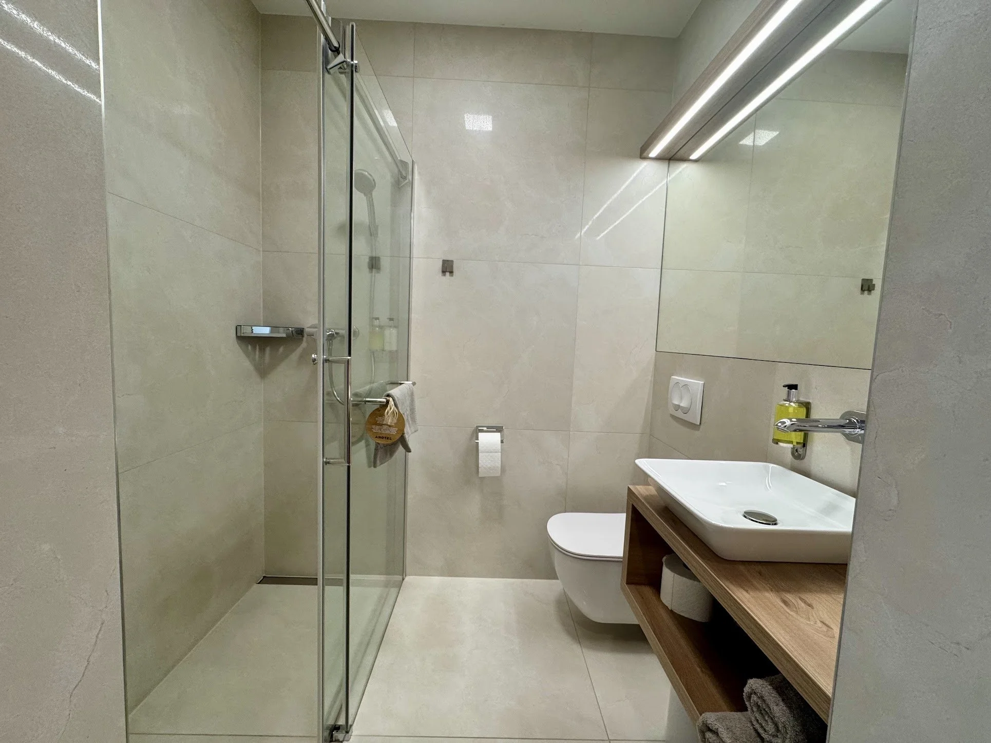 Modern bathroom with glass shower enclosure, white toilet, wooden vanity with white sink, mirror, and wall-mounted faucet, neutral tiled walls and floor, soap dispenser, and towels.