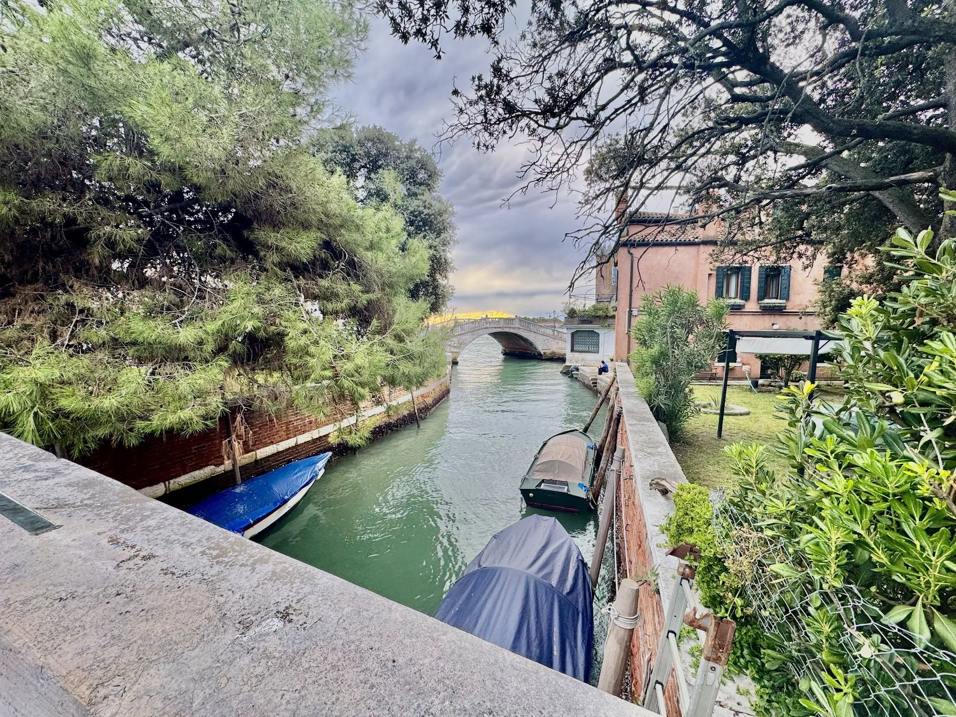 A canal scene with boats moored along the water, greenery on both sides, a stone bridge in the background, and a building with blue shutters on the right.