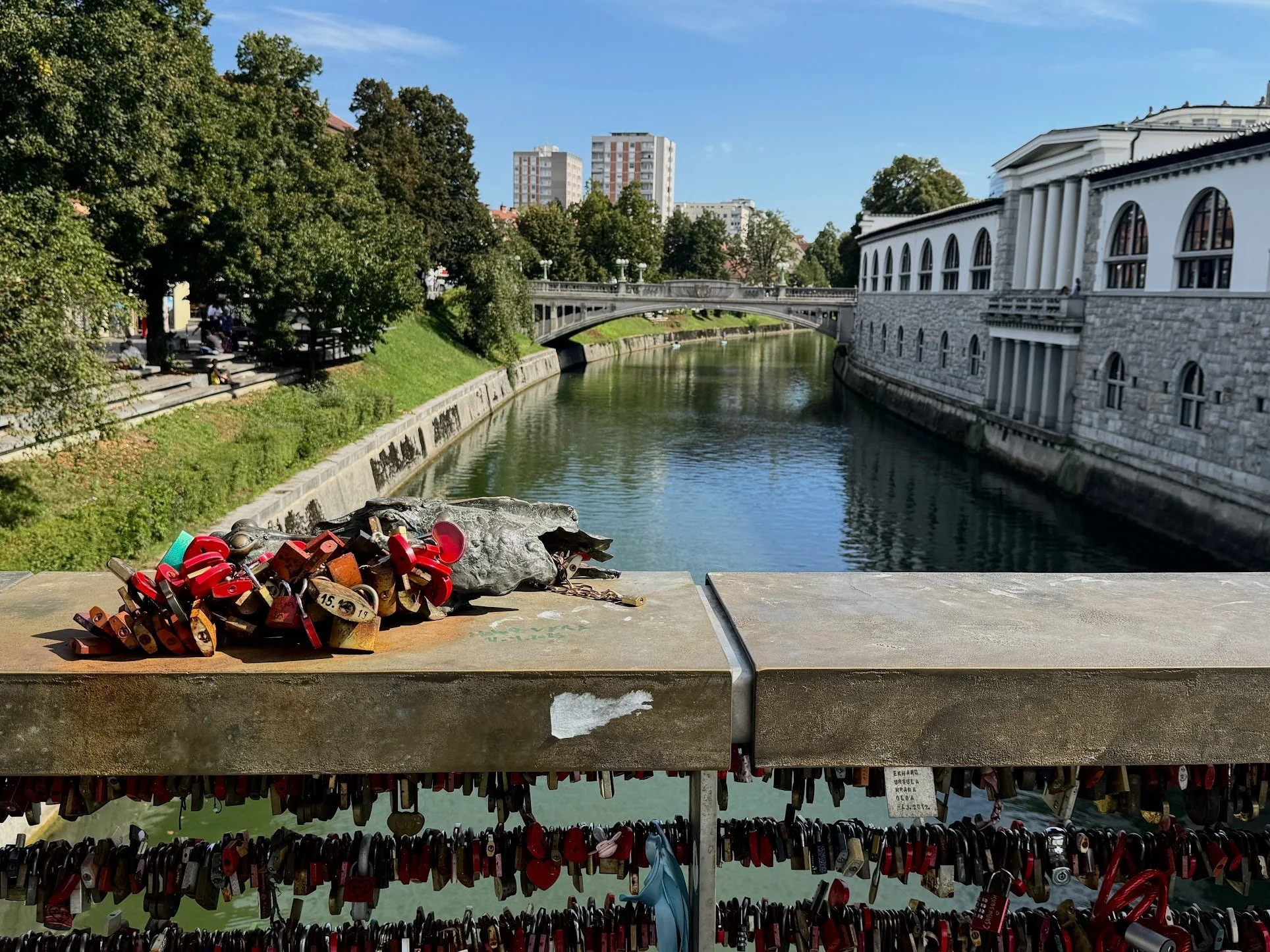 Bridge over a river with trees on the bank, and a building on the right side with classical columns and arched windows. In the foreground, a collection of love locks attached to a metal railing.