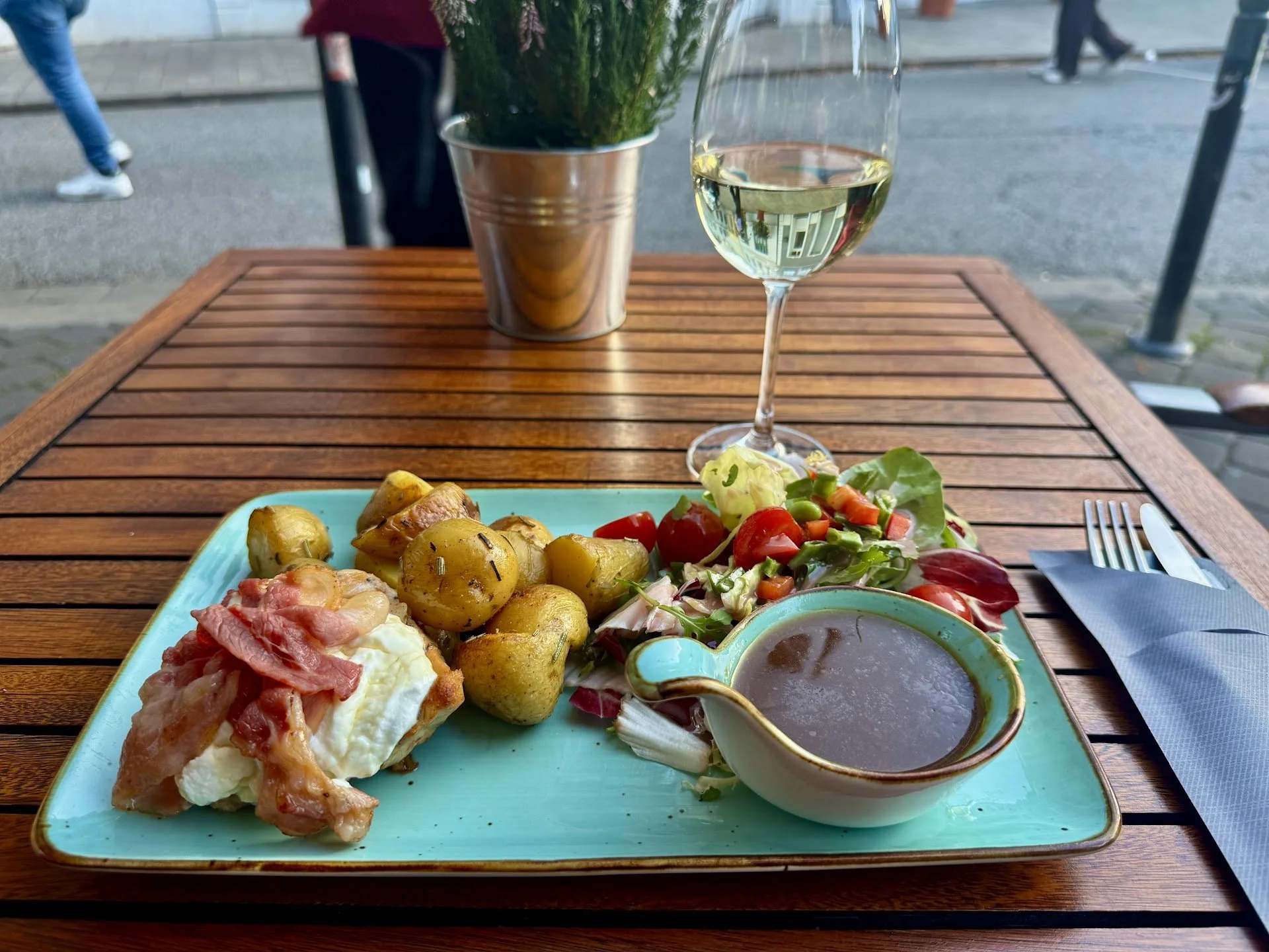 A plate of food with baked potatoes, ham, fried egg, salad, and a bowl of brown sauce, on a table outside. There is a glass of white wine and a plant next to it, with a blurred street scene in the background.