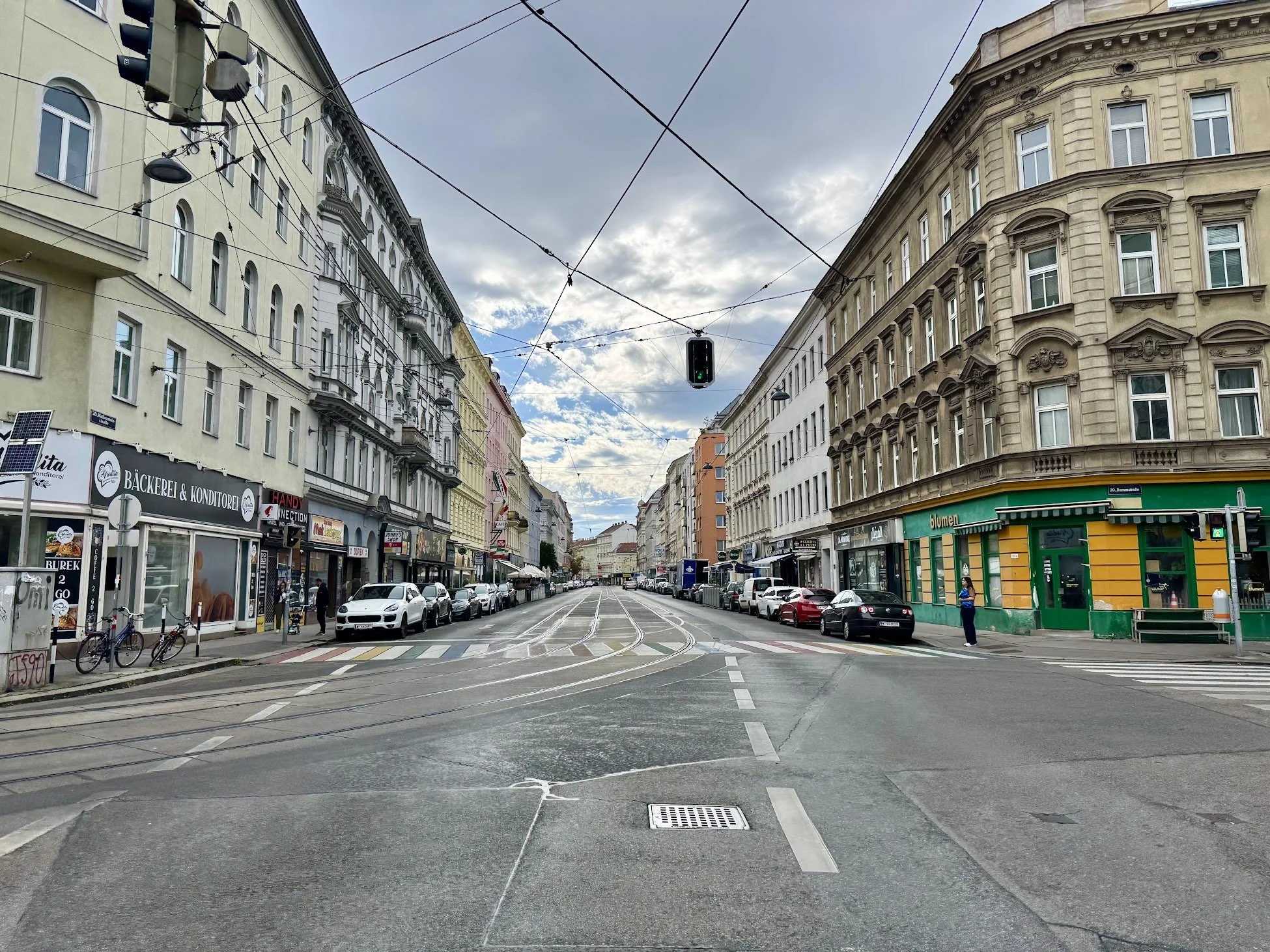 City street with historic European-style buildings on both sides, streetcar tracks in the middle, parked cars, and a traffic light hanging overhead. The sky is partly cloudy with some sunlight.