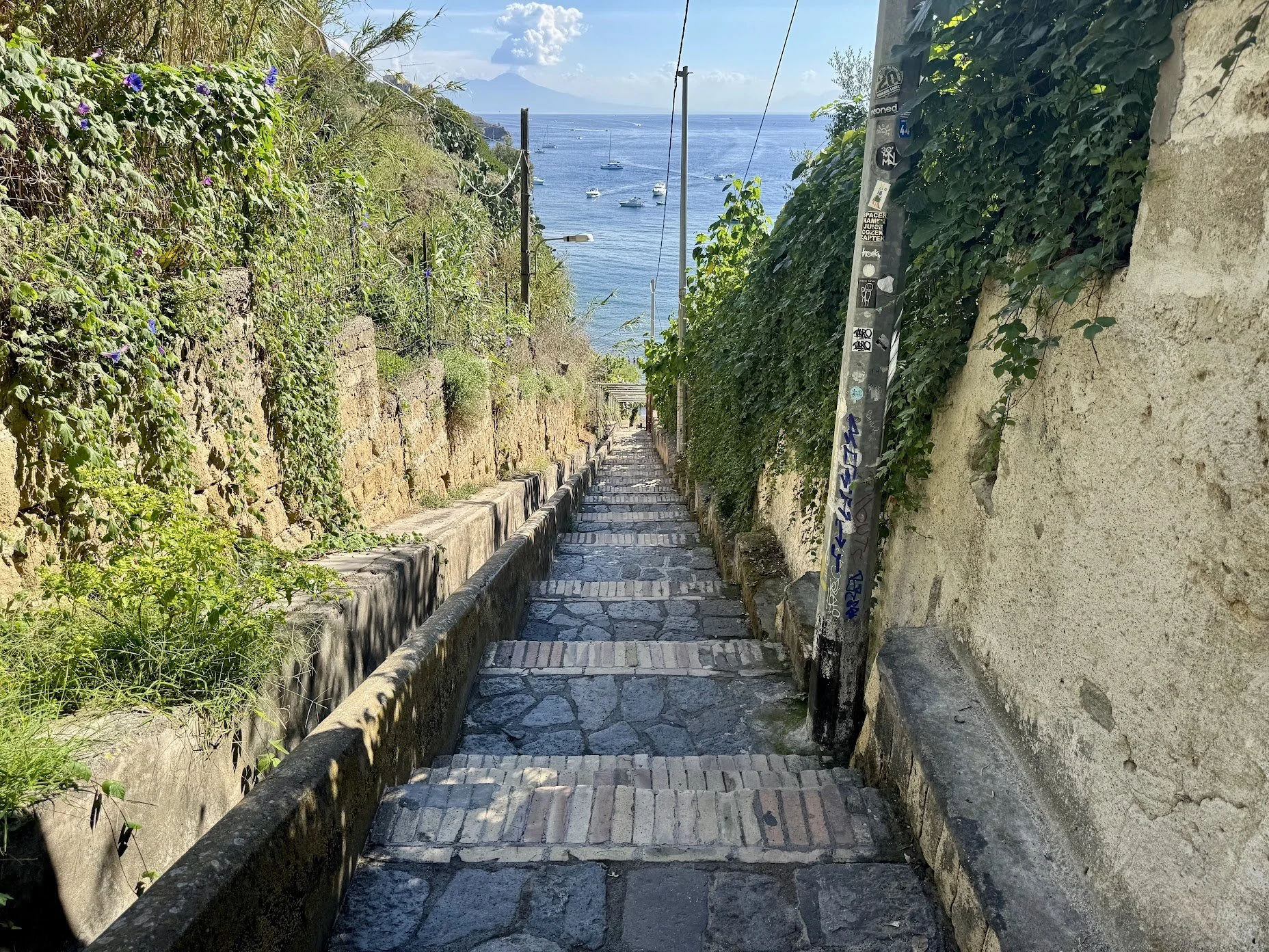 A narrow stone-paved pathway descending through a hillside alley with stone and brick walls, green foliage, and purple flowers on the left, and a stucco wall with green vines on the right. The background shows a view of a blue sea with several sailbo