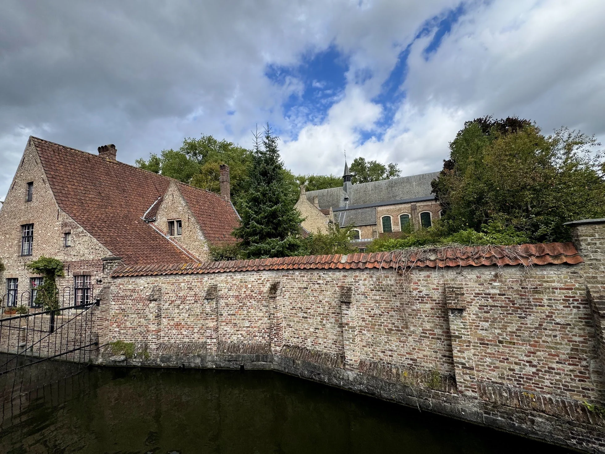 A brick building with a red tile roof and multiple chimneys, surrounded by a brick wall, trees, and a small waterway in the foreground under a partly cloudy sky.