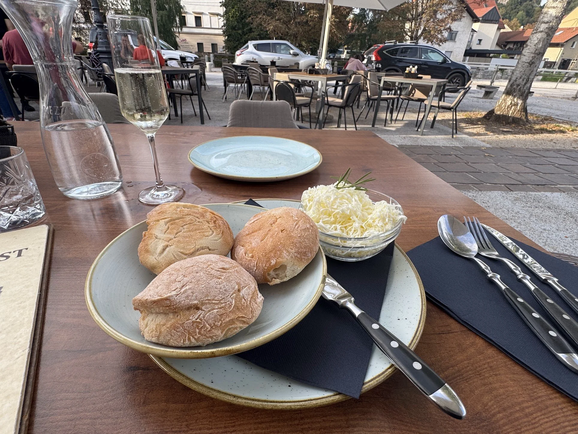 Breakfast served outdoors on a wooden table, featuring three bread rolls, a small bowl of shredded cheese, a glass of sparkling wine, a pitcher of water, and a glass of water, with empty plates and utensils, in a restaurant patio with outdoor seating