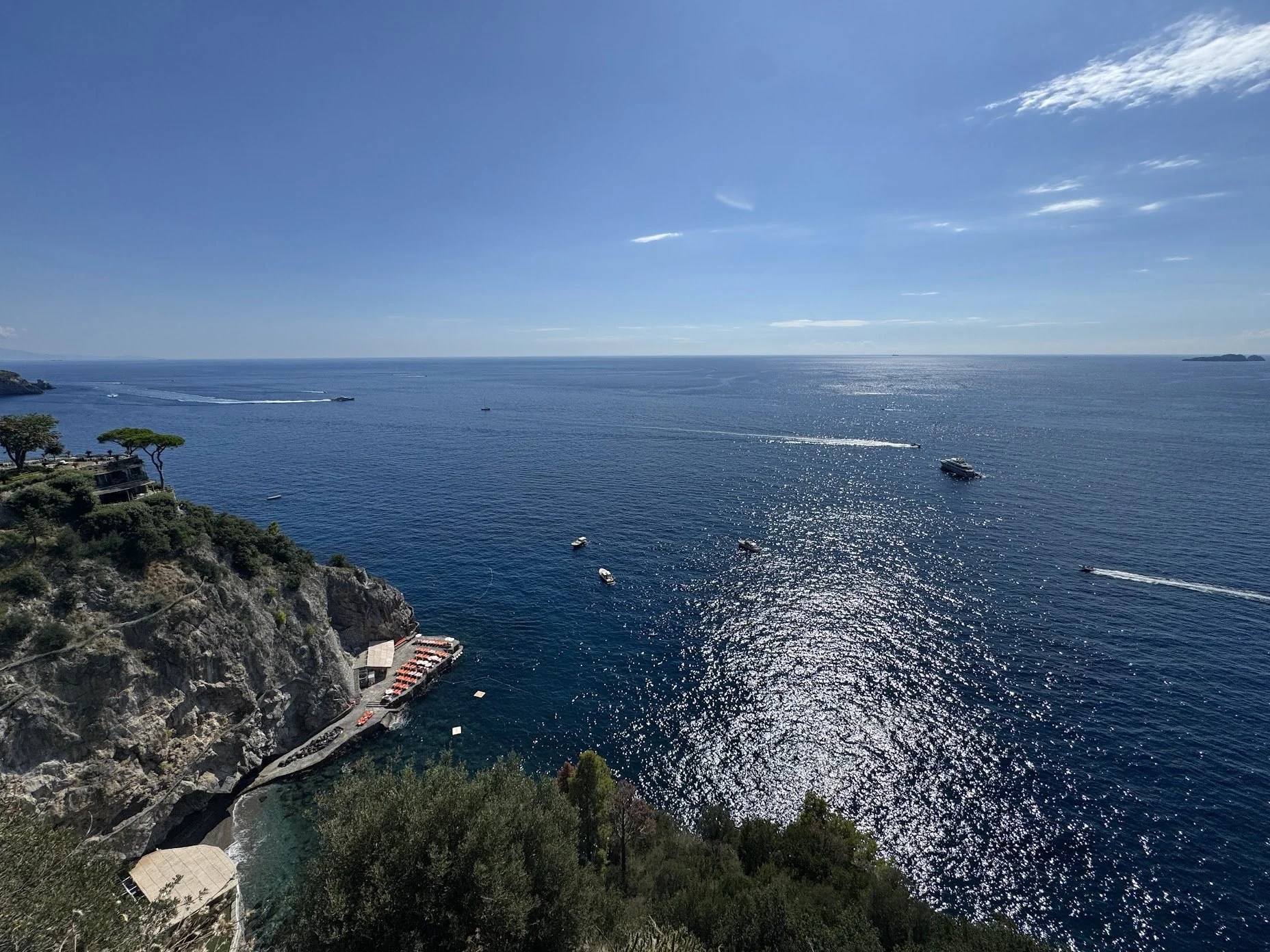 A view of a large body of water with several boats and speedboats creating wakes, seen from a rocky hillside with trees and buildings at the bottom left corner, under a mostly clear blue sky with a few clouds.