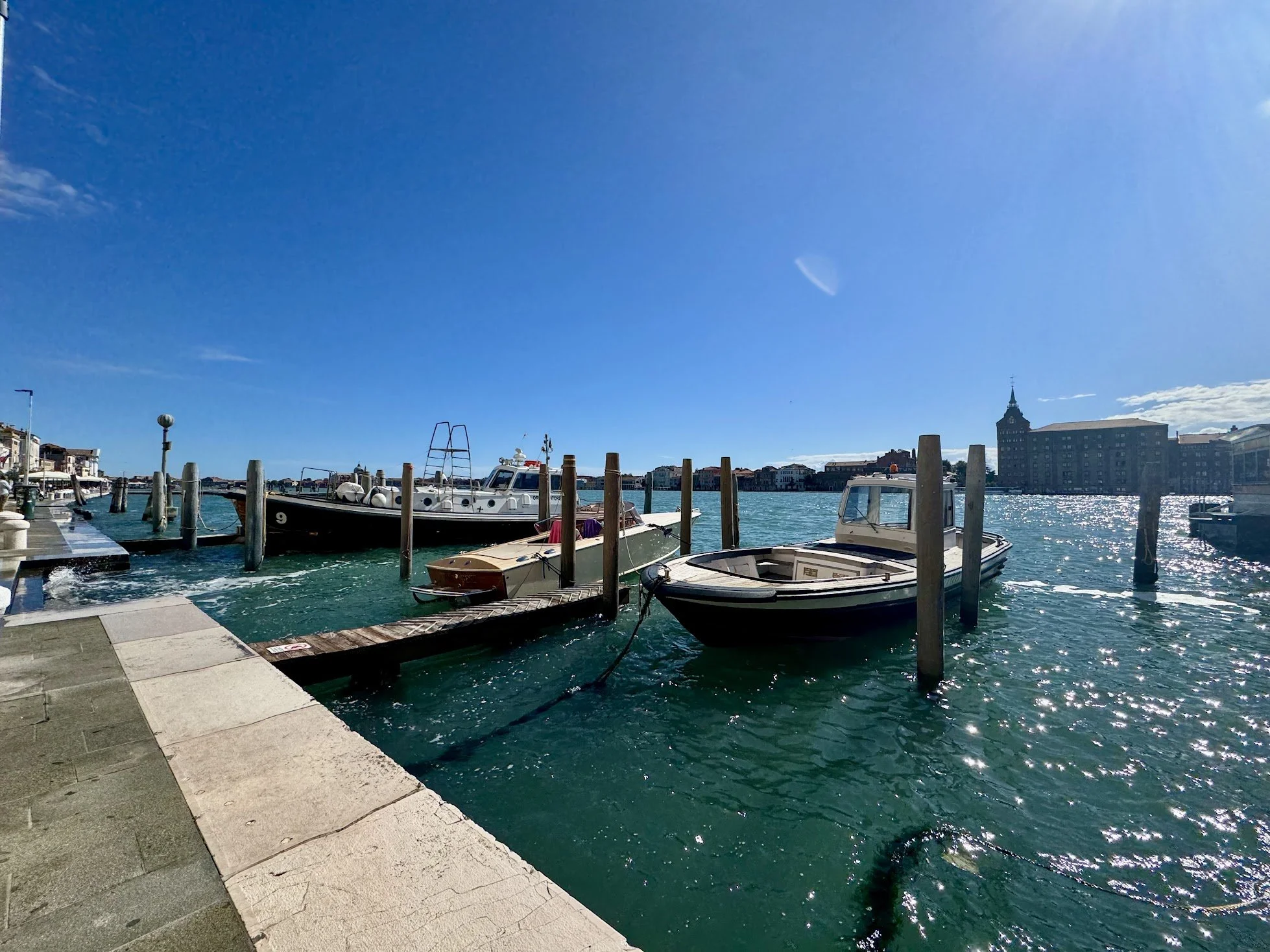 Boats docked at a marina on a sunny day with clear blue sky, across the water from a city skyline with historic and modern buildings.