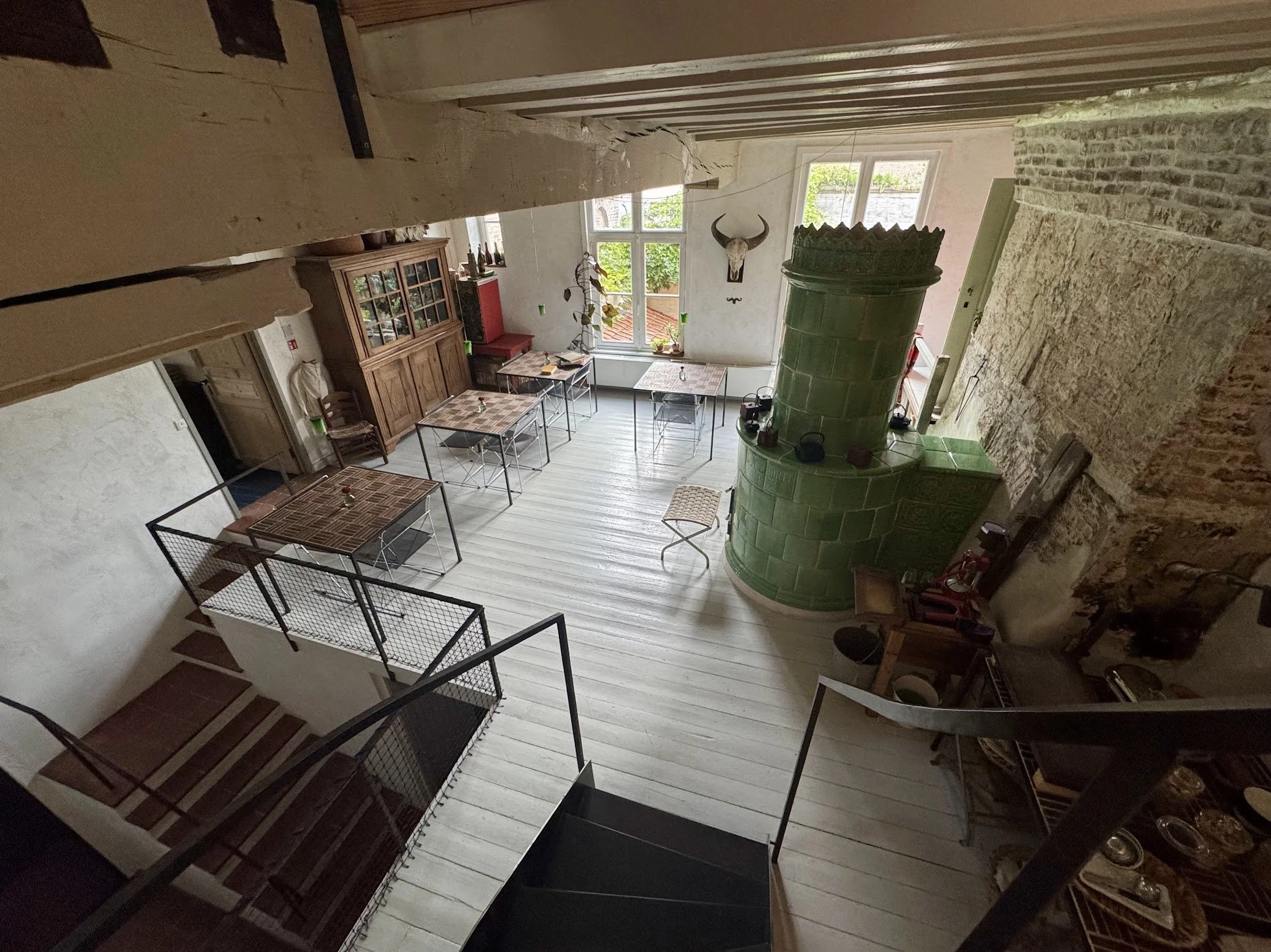 A rustic dining area with wooden floors and white walls, featuring a green tiled oven, large windows, and decor including cattle skulls with horns