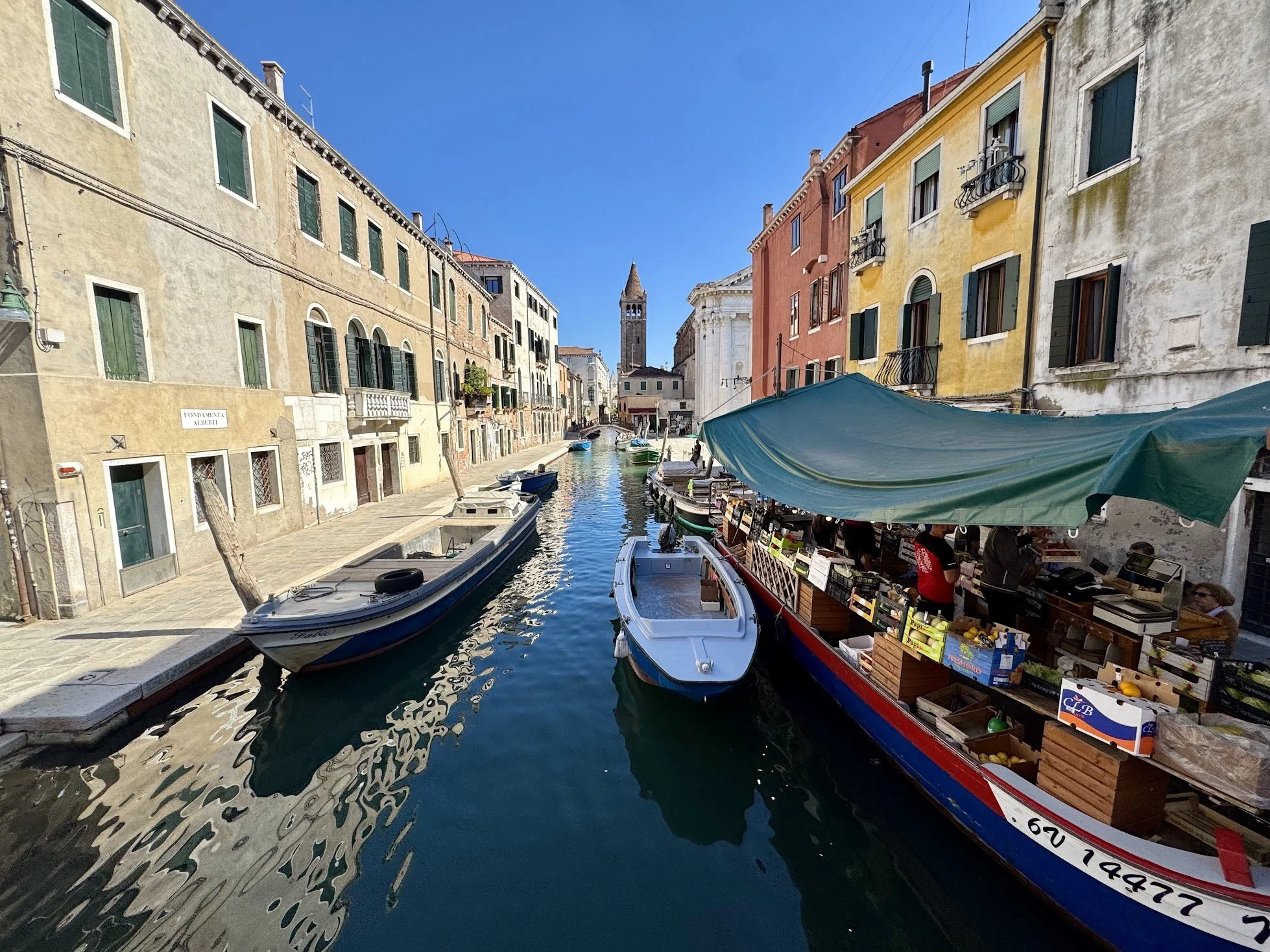 A canal scene in Venice with boats and colorful buildings lining the waterway, a clock tower in the background, and market stalls with umbrellas along the canal.