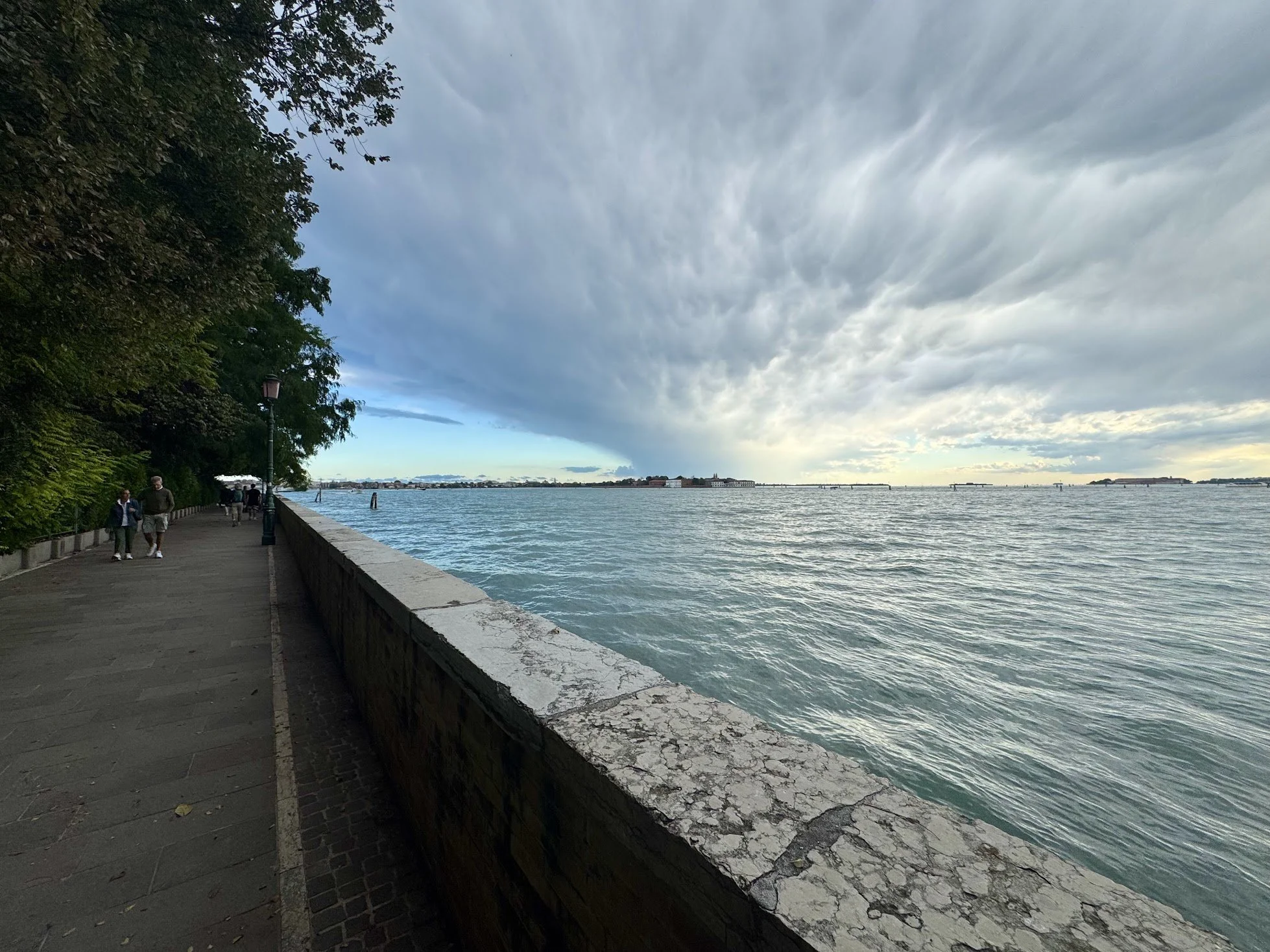 A lakeside promenade with a few people walking, a lamp post, trees on the left, and a broad water body under cloudy skies.