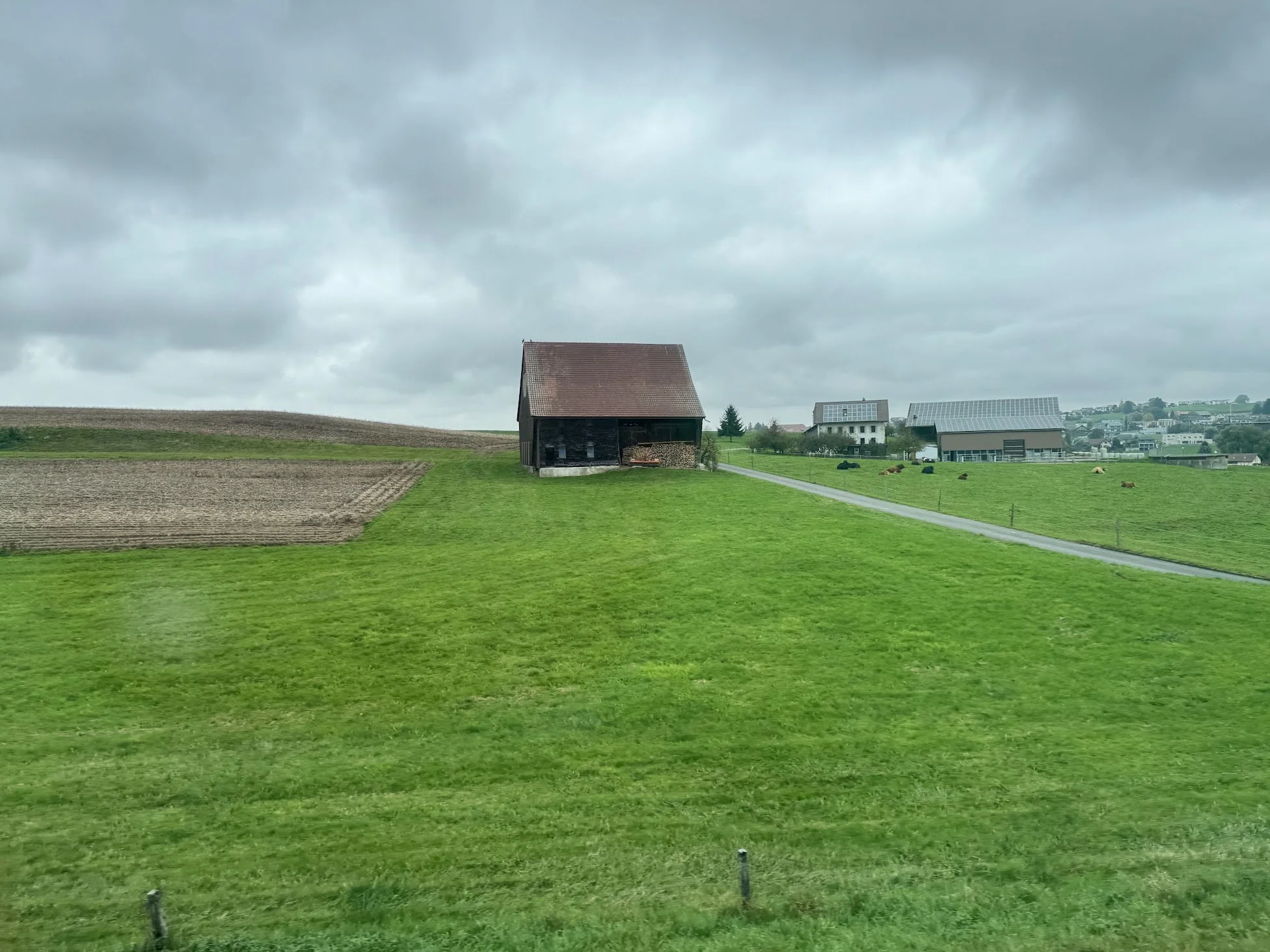 A rural landscape with green fields, a farm building with a red roof, and several houses in the background under a cloudy sky.